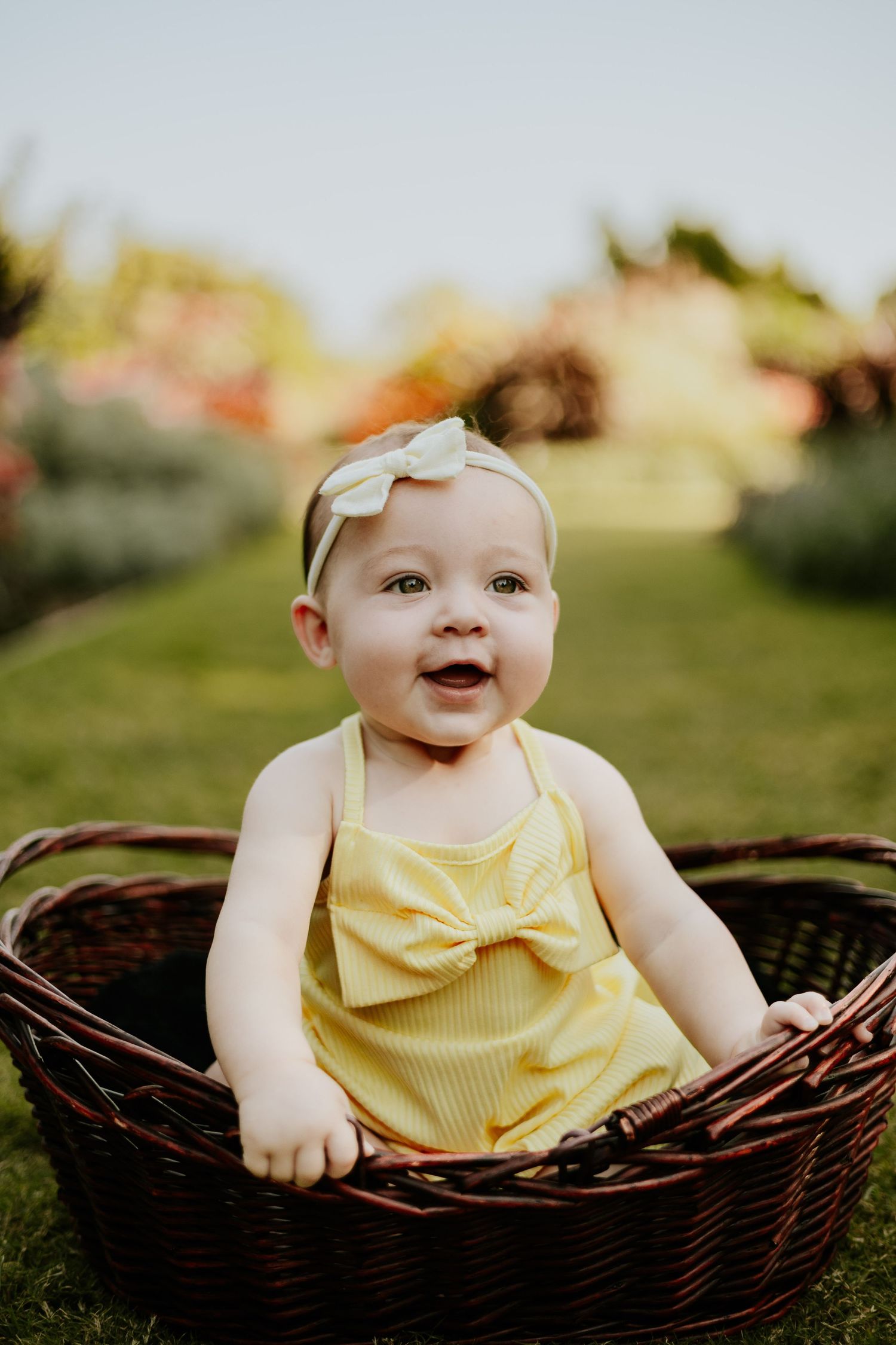 Happy baby girl in yellow bow romper and white headband smiling in a wicker basket outside.