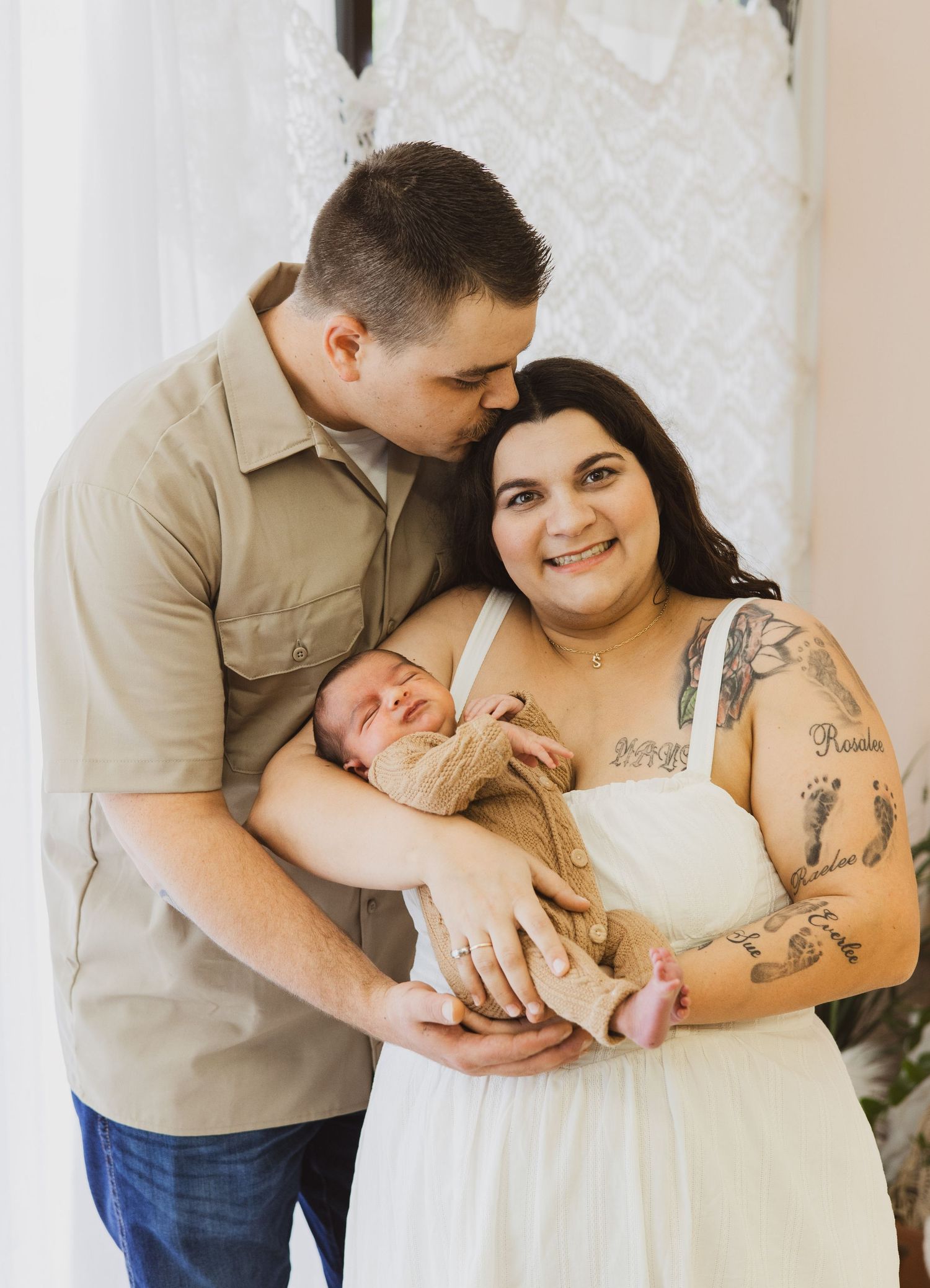 Smiling tattooed mother in white dress and father in tan shirt holding newborn baby together.