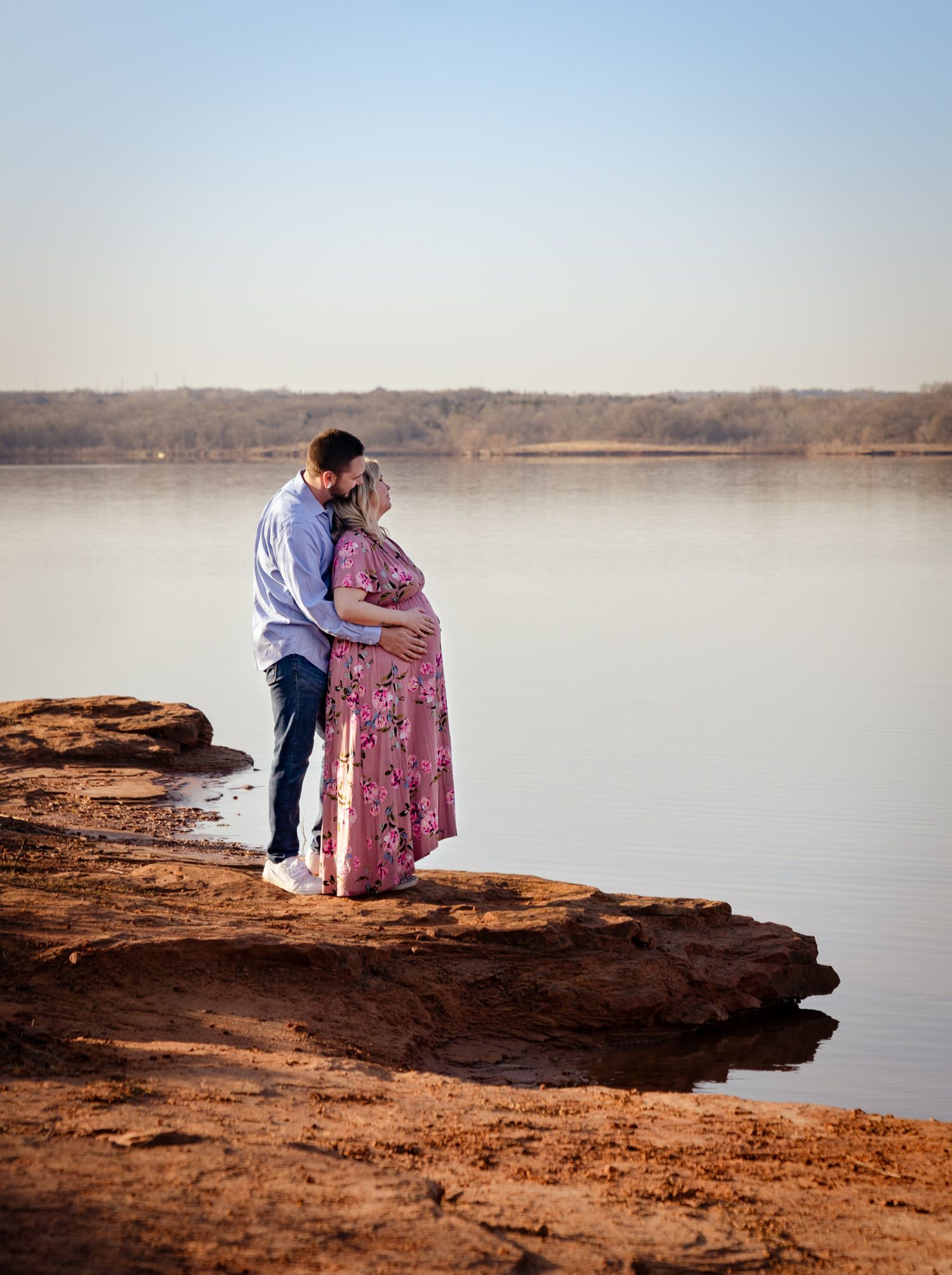 Couple sharing a tender kiss by a serene lake at golden hour during a maternity photo session.