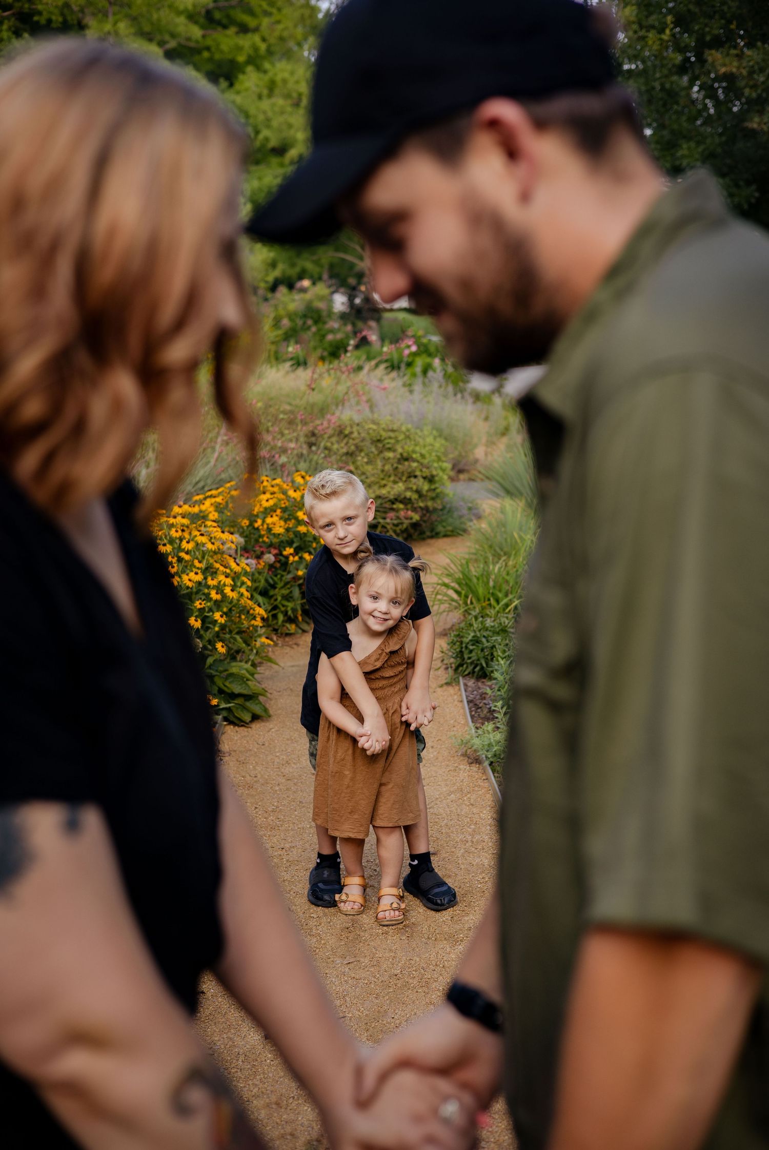 Two young siblings hugging on garden path while parents hold hands in soft-focus foreground.