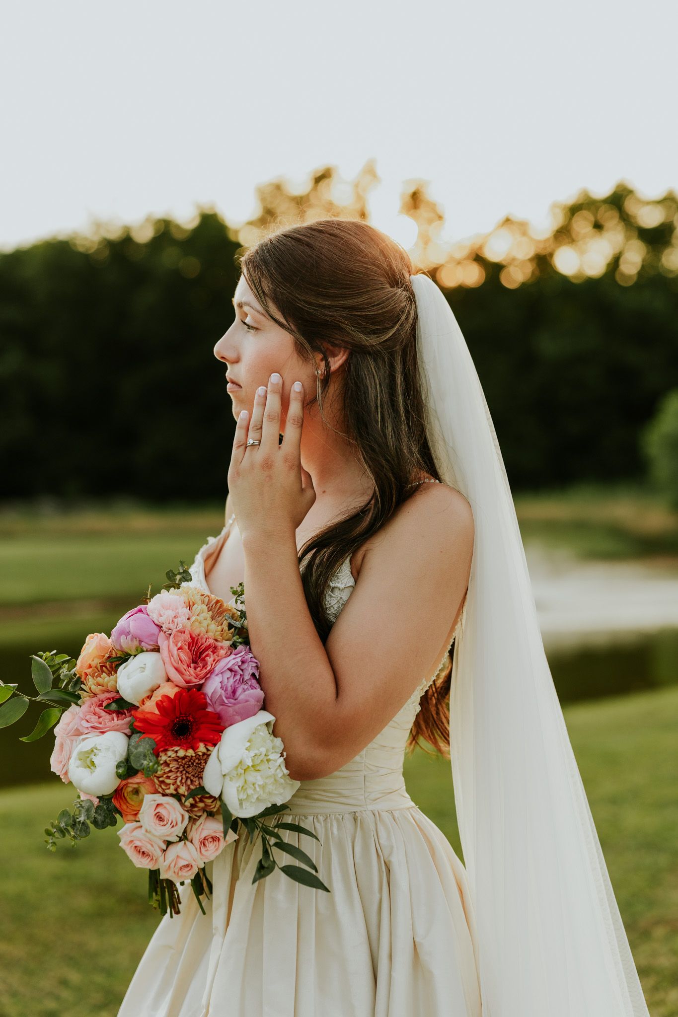 Emotional bride in white gown and veil holds colorful bouquet, hand to face, at golden hour outdoor wedding.