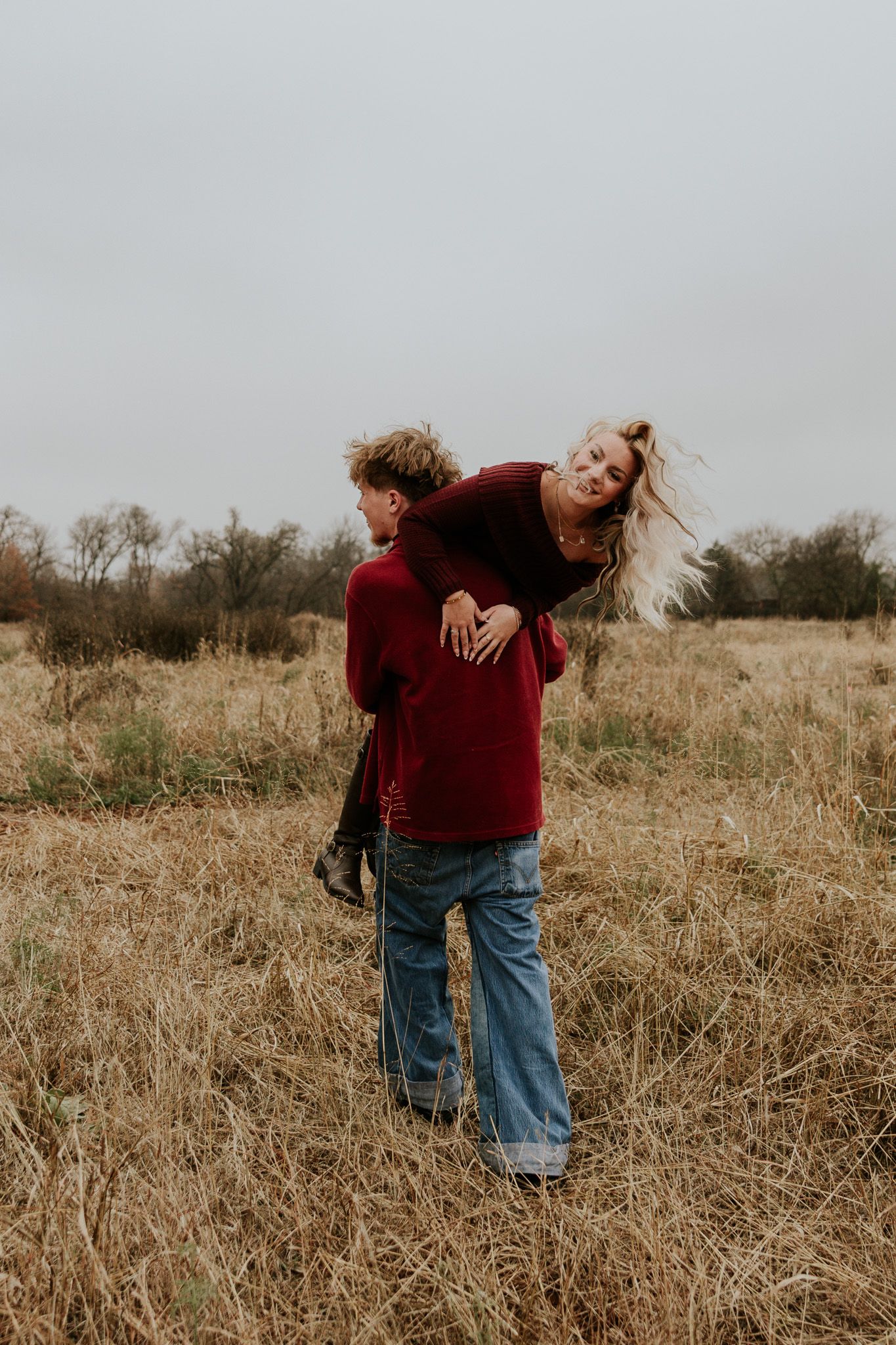 Man giving woman a piggyback ride in a golden field, both wearing matching burgundy sweaters.