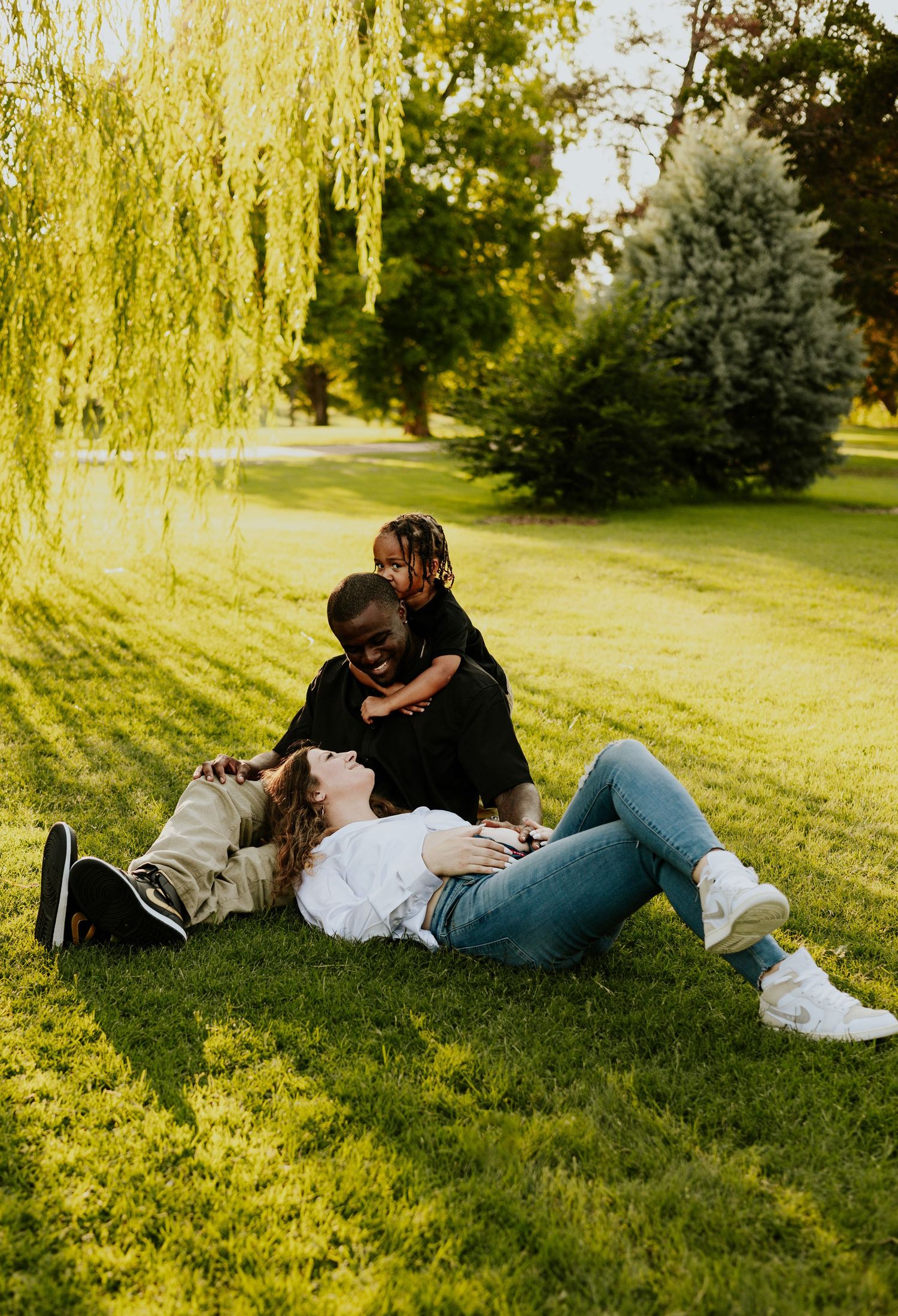 Happy interracial family relaxing on green grass under willow tree with child climbing on dad.