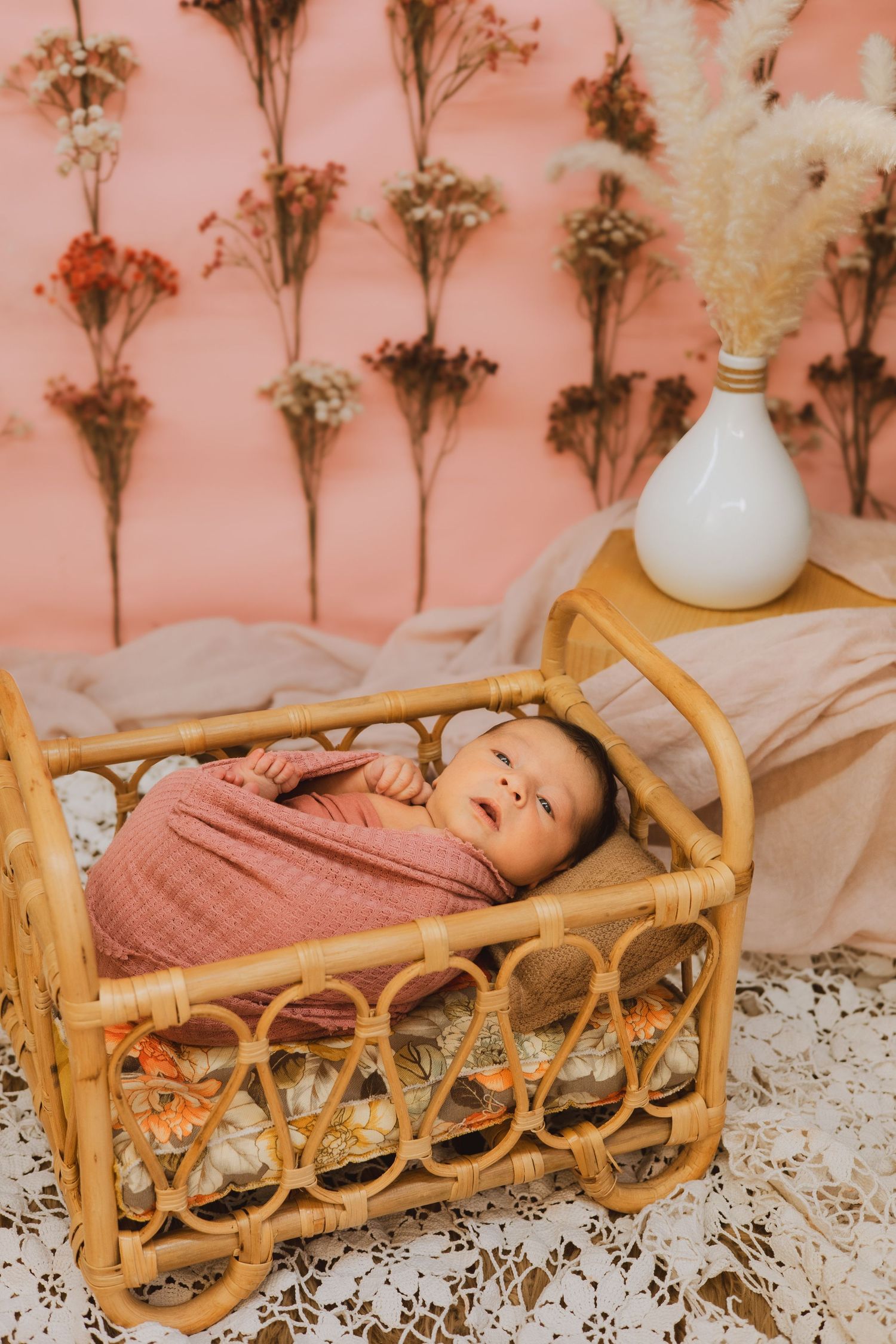 Newborn baby swaddled in pink wrap lying in a rattan crib against a pink floral wall with dried flowers.