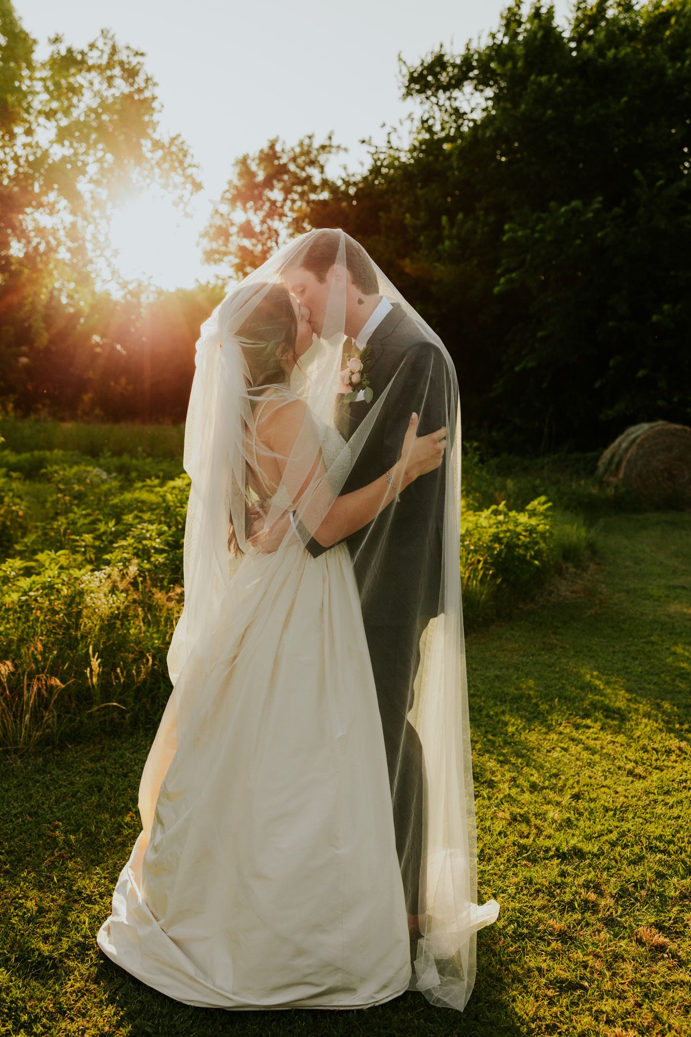 Bride and groom sharing a kiss under a veil at golden hour in a lush garden setting.