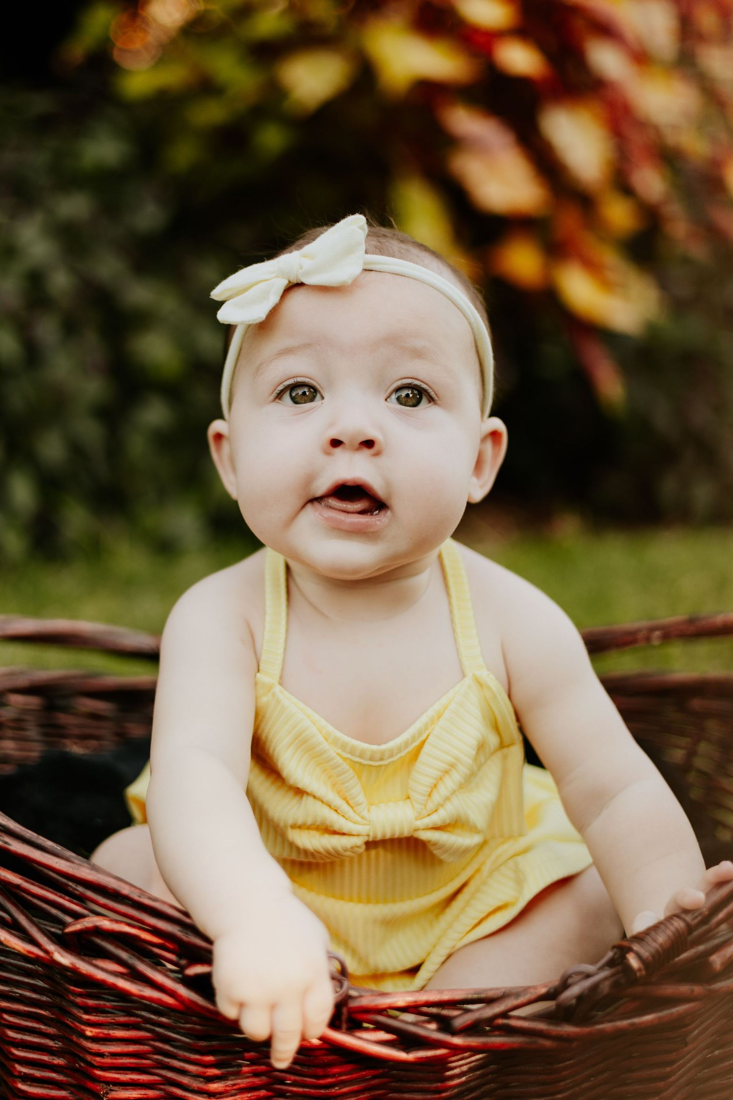 Sweet baby girl in yellow romper and white bow headband sitting in a wicker basket in garden.