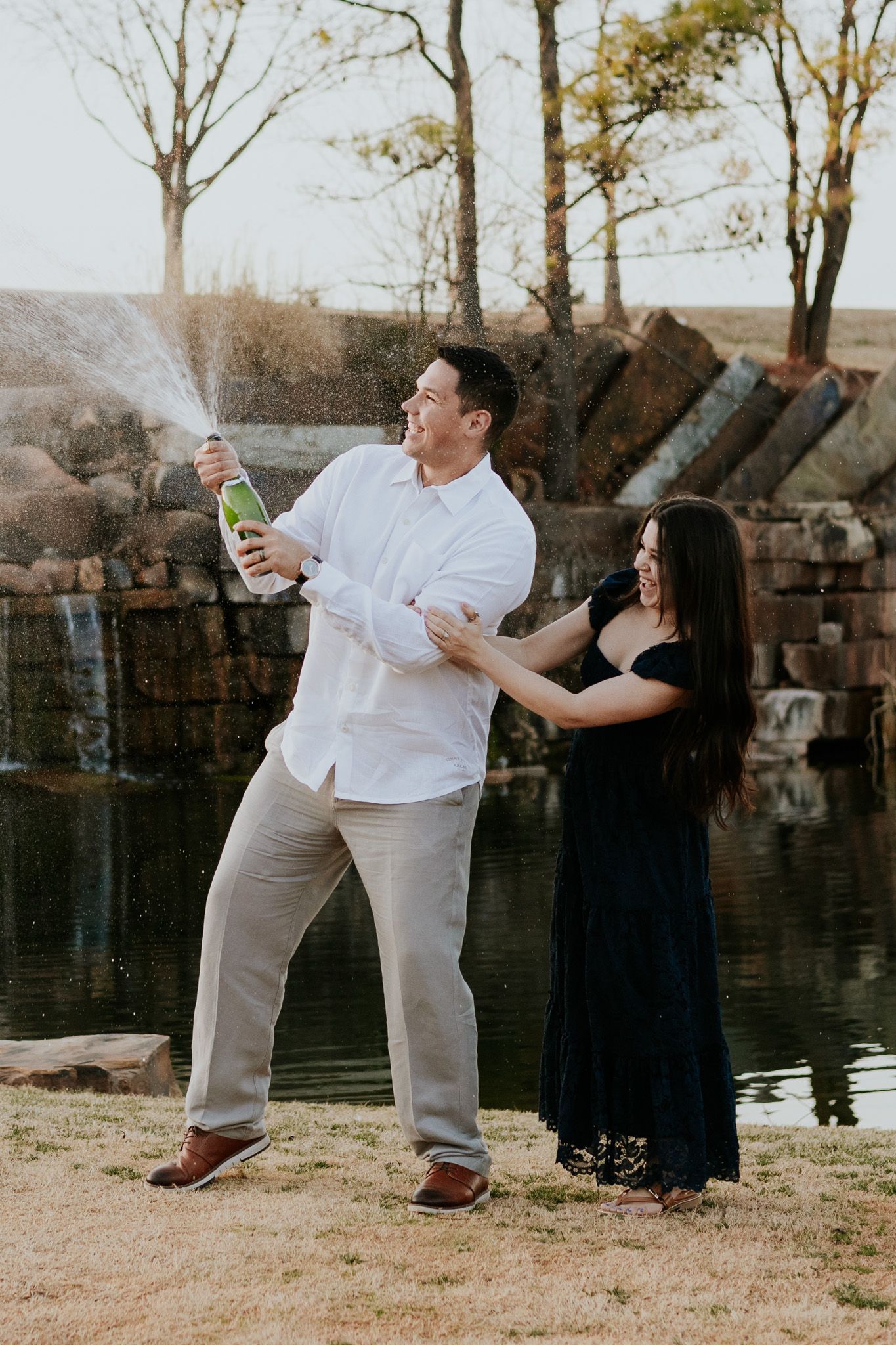Joyful couple popping champagne bottle together near stone waterfall, man in white shirt and woman in black dress.