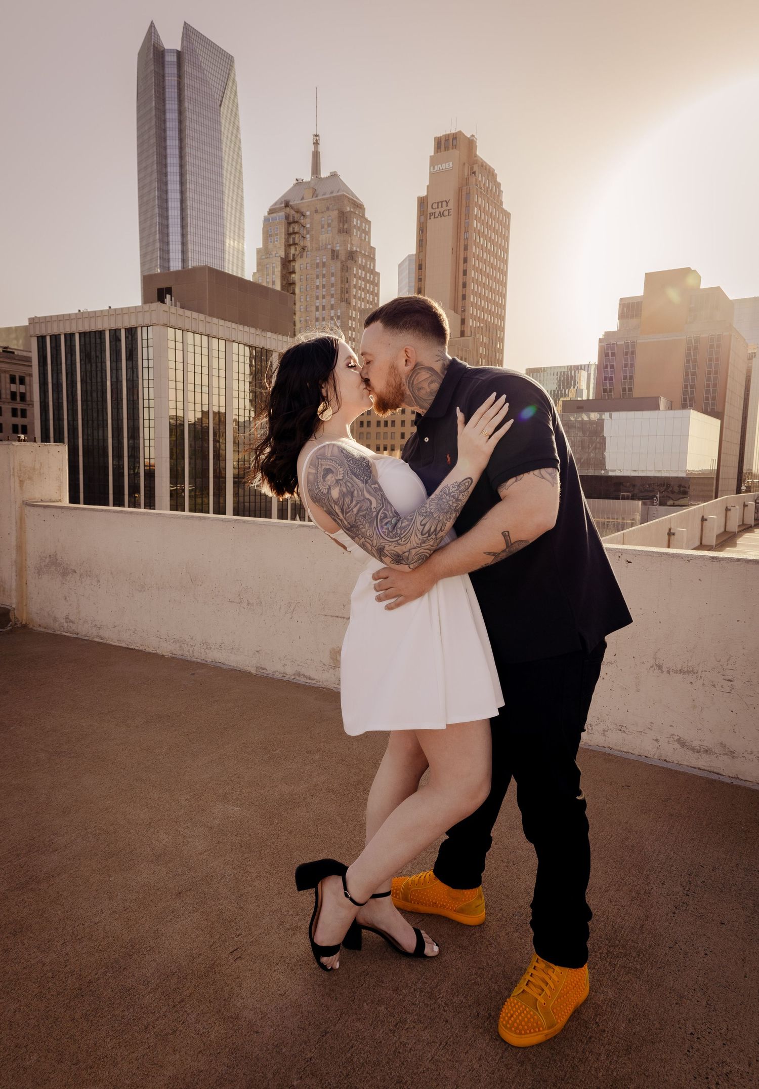 Tattooed couple kissing in white dress on rooftop at sunset.