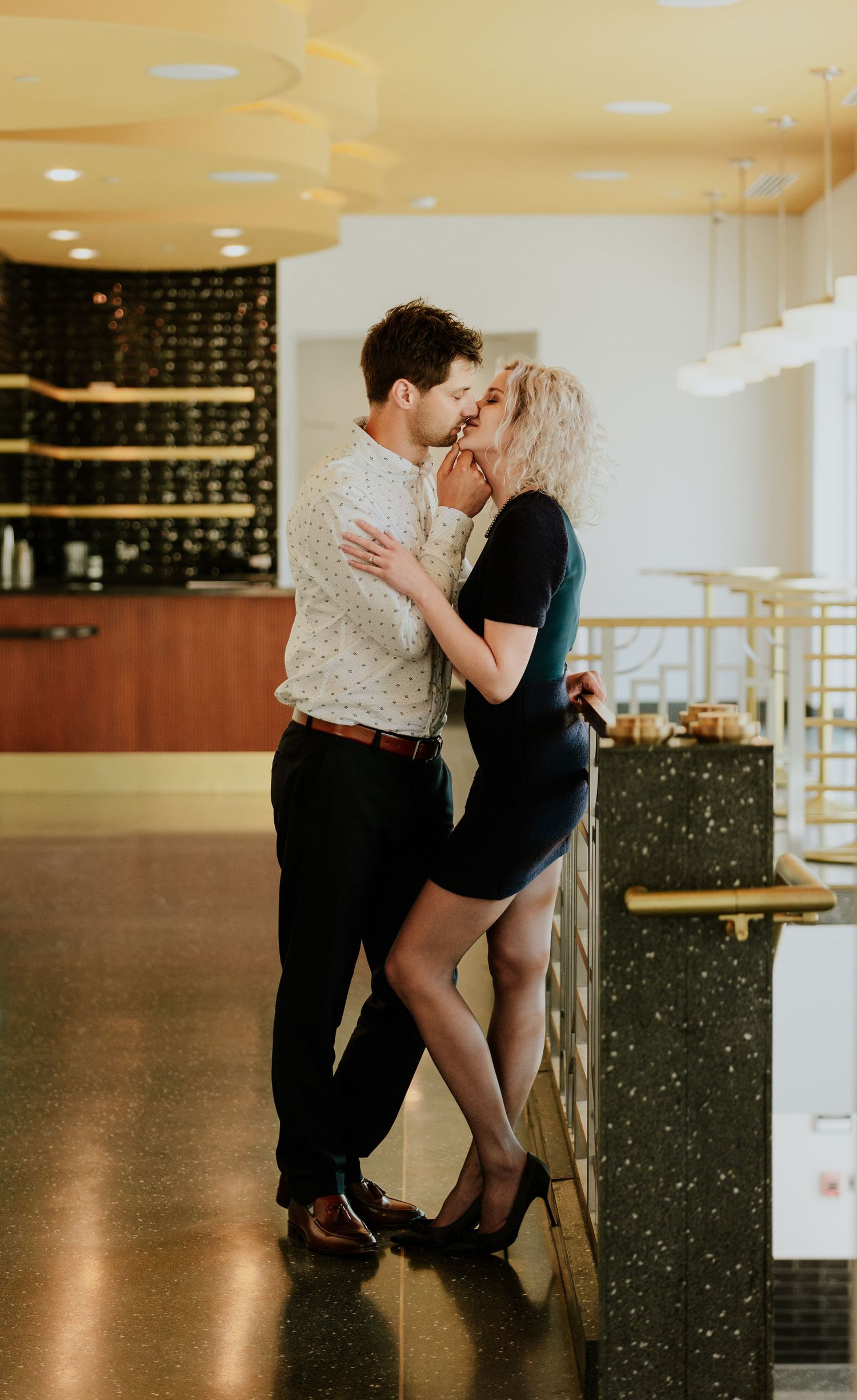 Couple sharing a kiss near a bar counter, woman in black mini dress and heels, man in dress shirt.