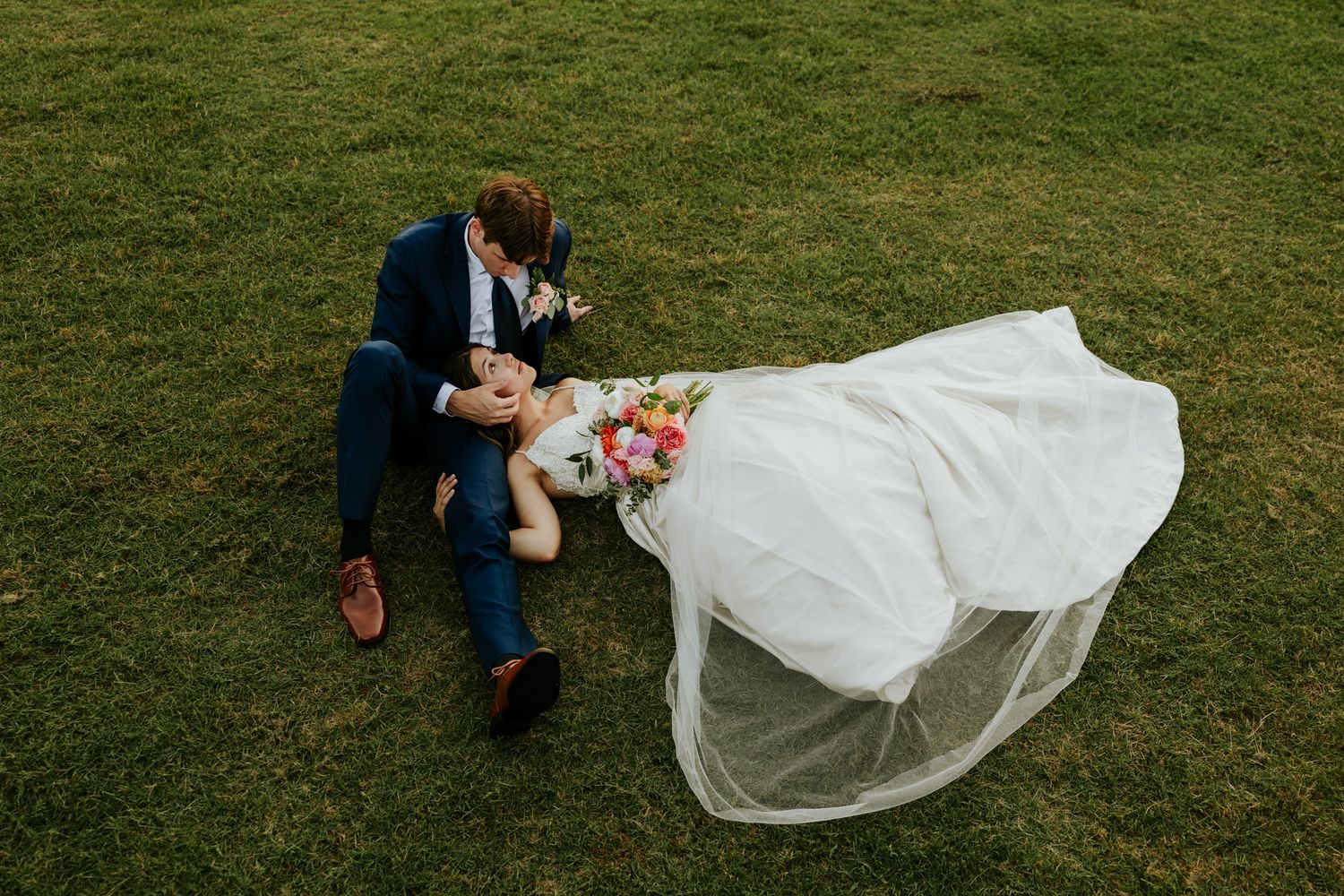 Aerial view of bride and groom sitting on green grass, bride in white gown with flowing veil and colorful bouquet.