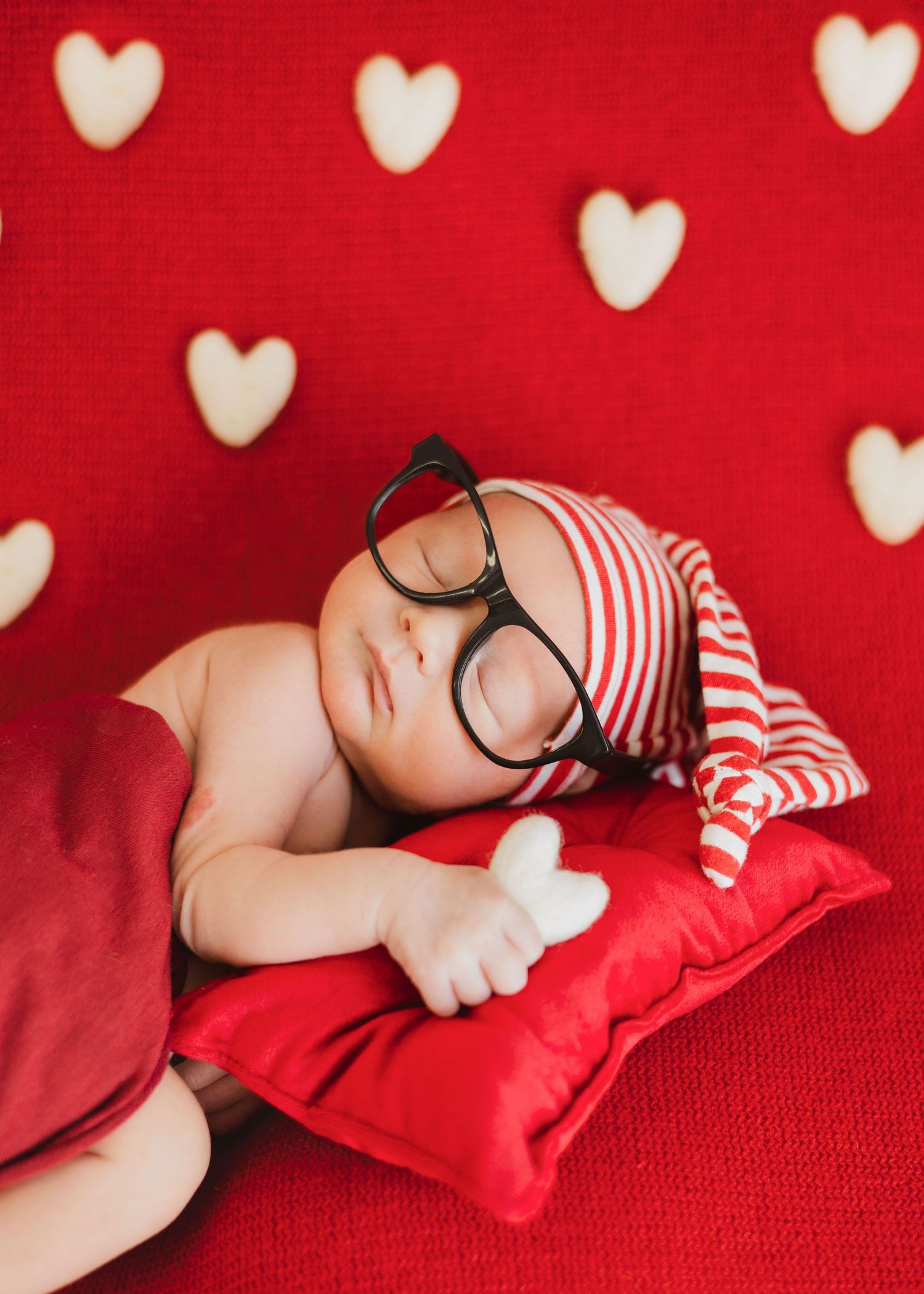 Adorable sleeping newborn wearing oversized glasses and striped hat on red heart-patterned Valentine backdrop.