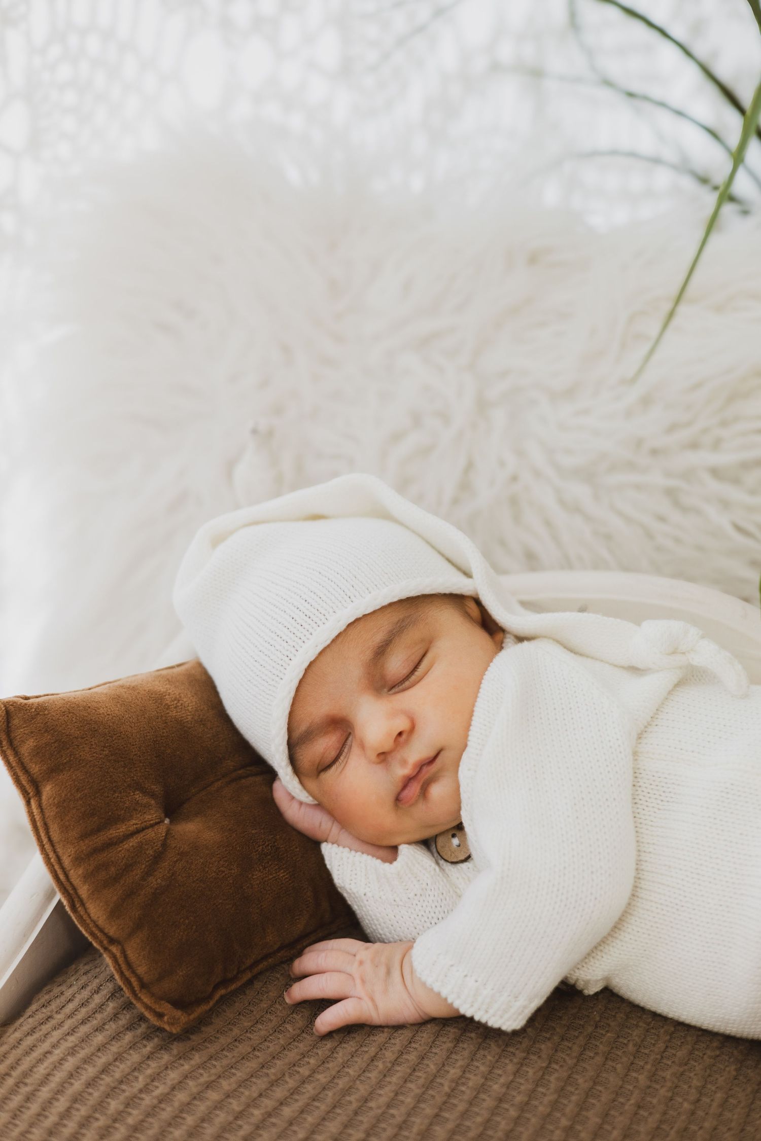 Sleeping newborn baby in white knit outfit and hat resting on brown velvet pillow.