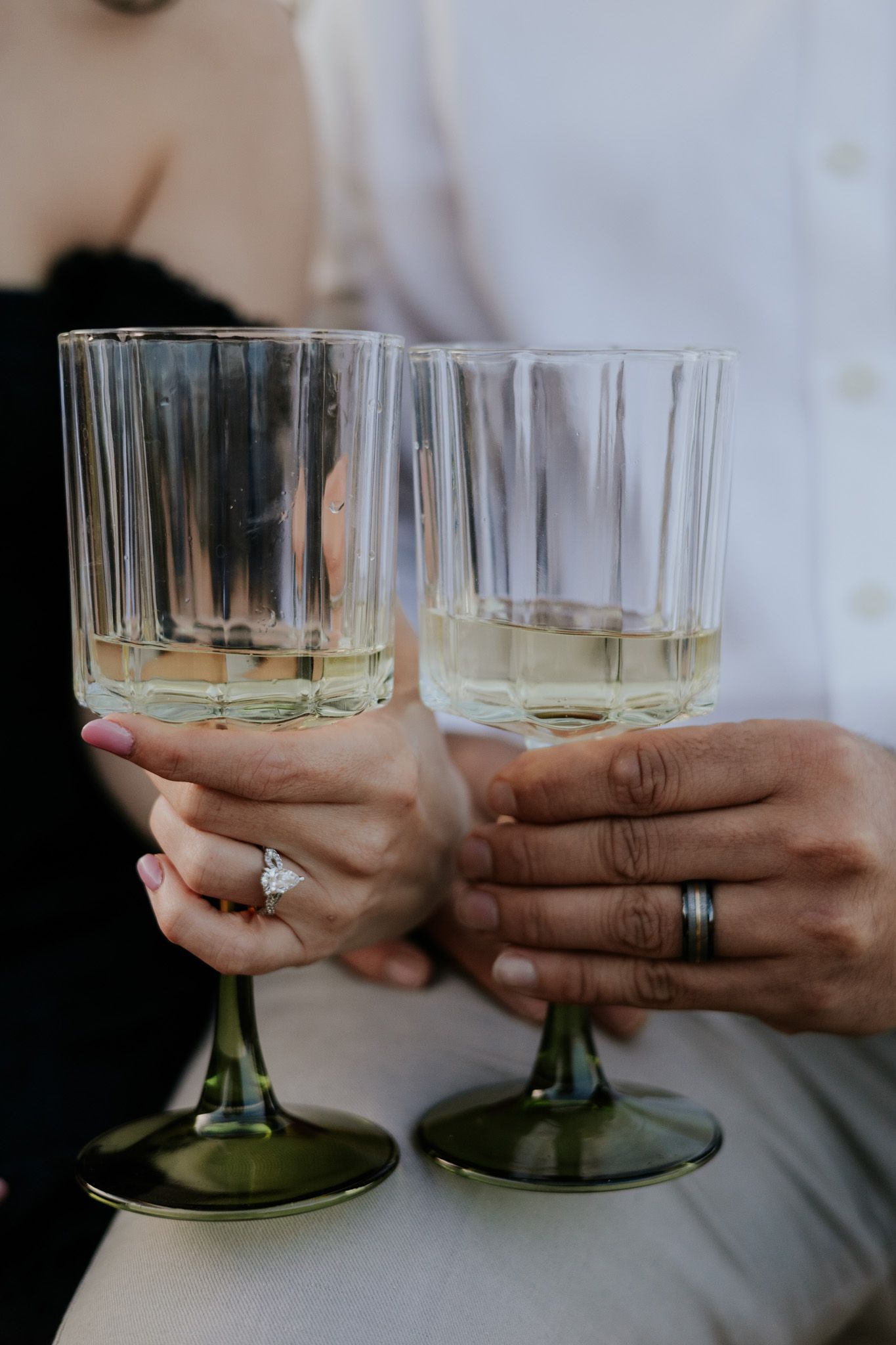Close-up of couple holding elegant green-stemmed champagne glasses, woman wearing diamond engagement ring.