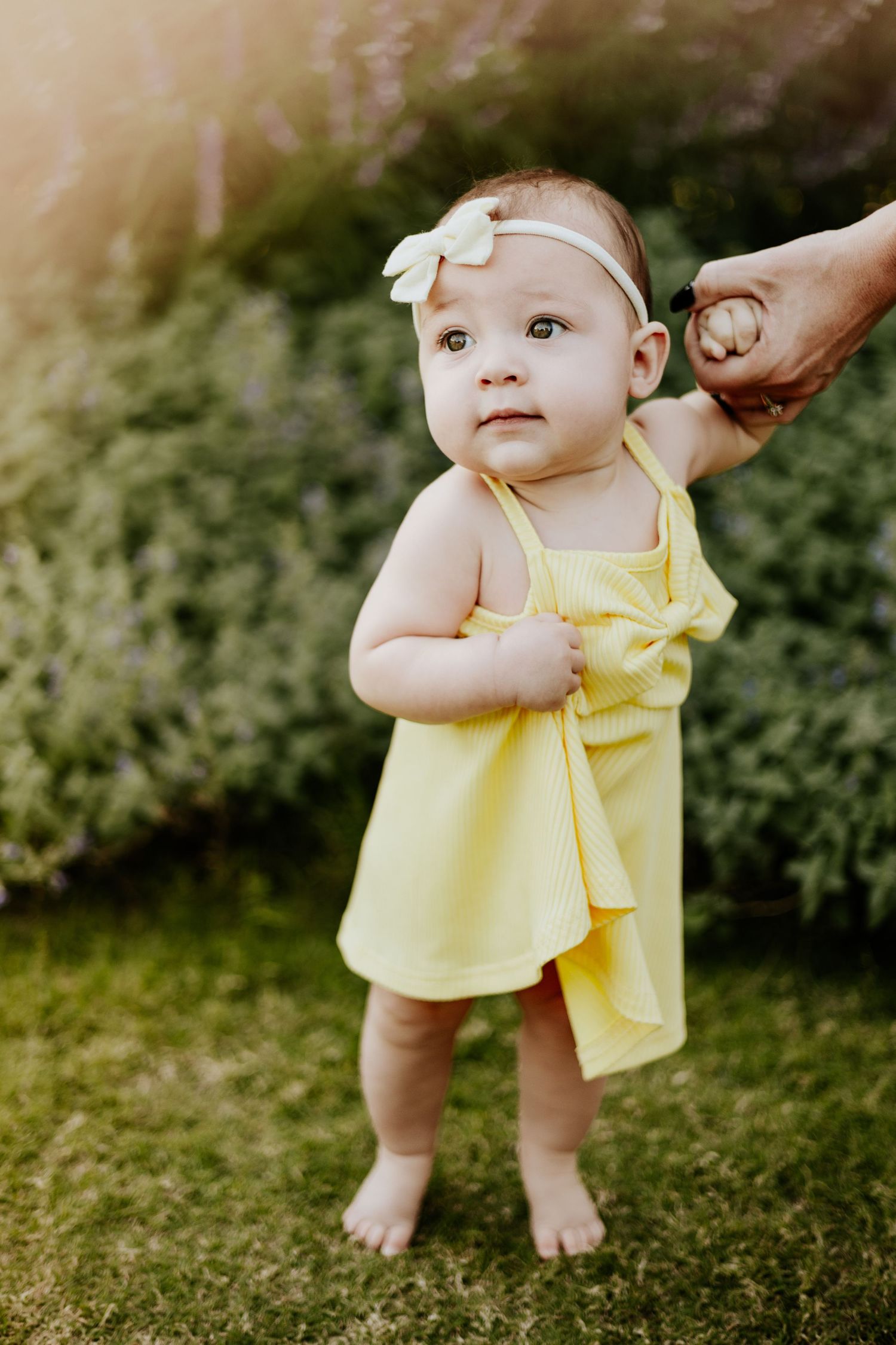 Baby girl in yellow dress and bow headband standing barefoot in garden holding an adult hand.