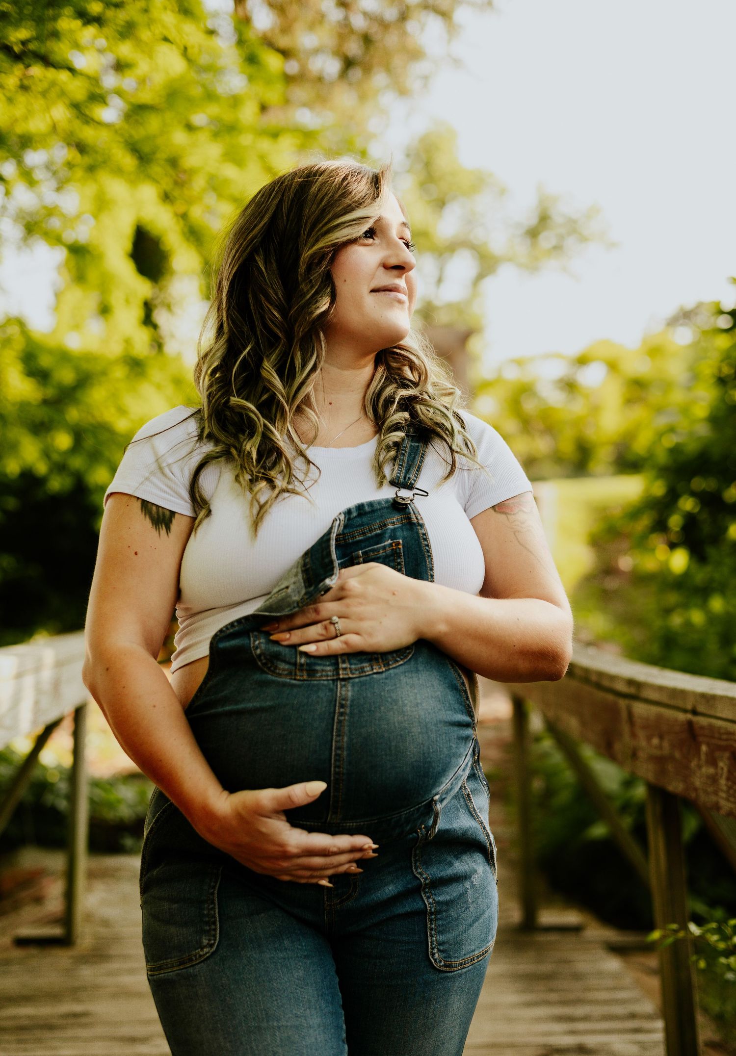 Pregnant woman in denim overalls and white tee cradling baby bump outdoors on wooden bridge surrounded by green trees.