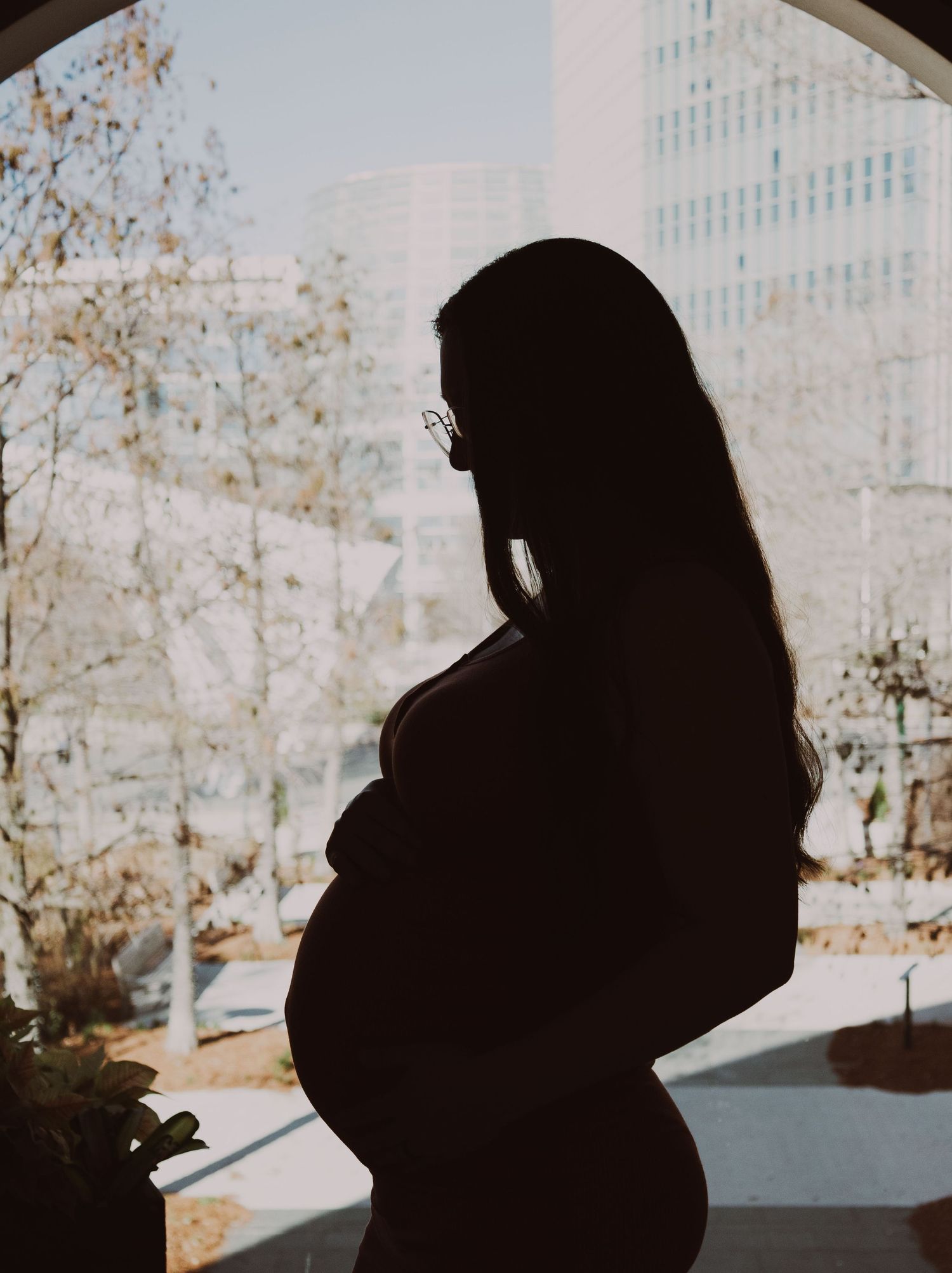 Silhouette of a pregnant woman with long hair standing by a large window in an urban setting.
