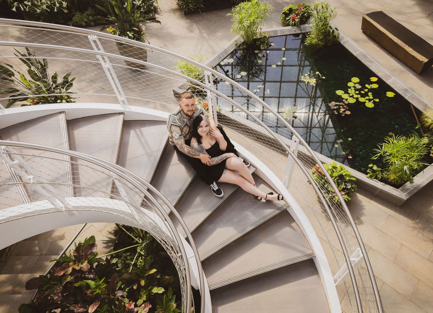 Couple sitting together on spiral staircase overlooking lush garden atrium.