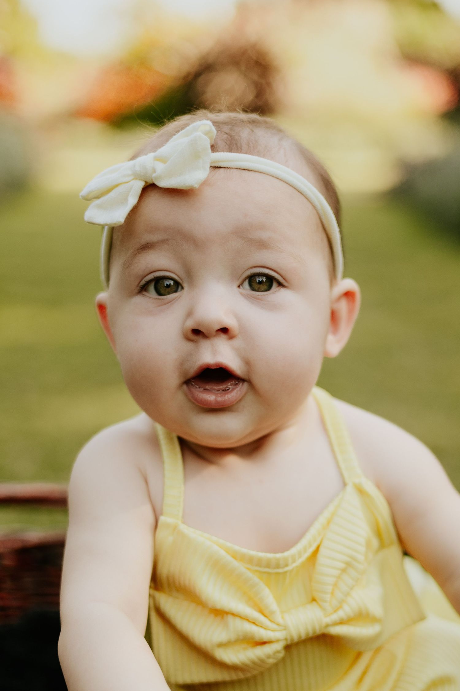 Close-up of cute baby girl in yellow dress and bow headband with surprised expression outdoors.