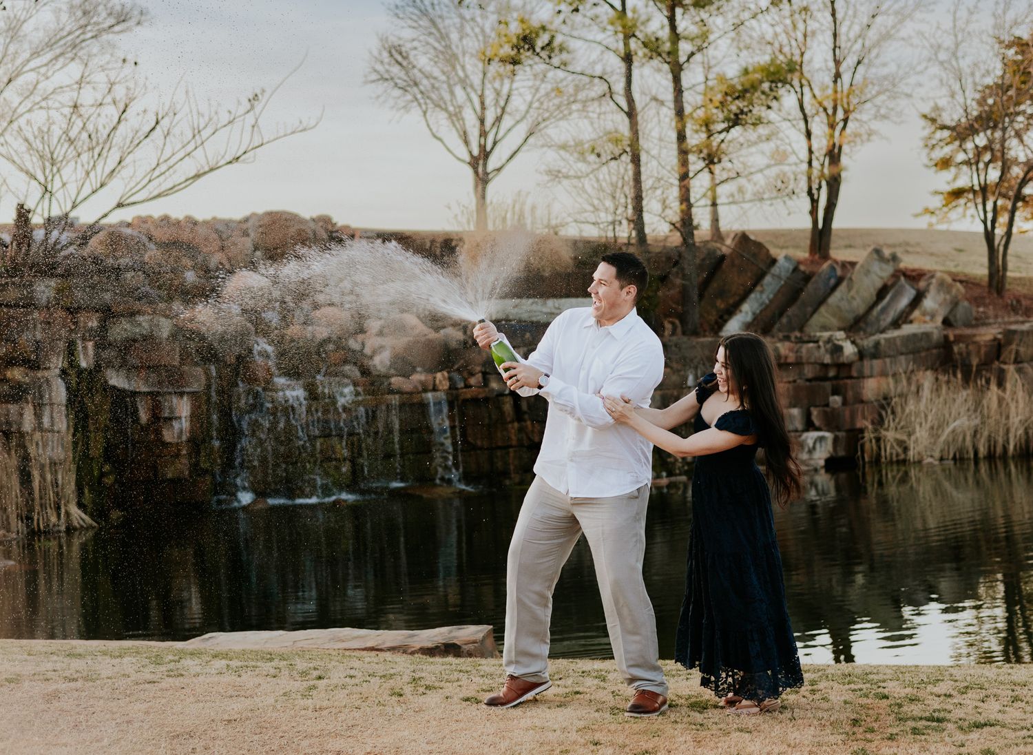 Couple celebrating engagement by popping champagne bottle near scenic waterfall and pond outdoors.