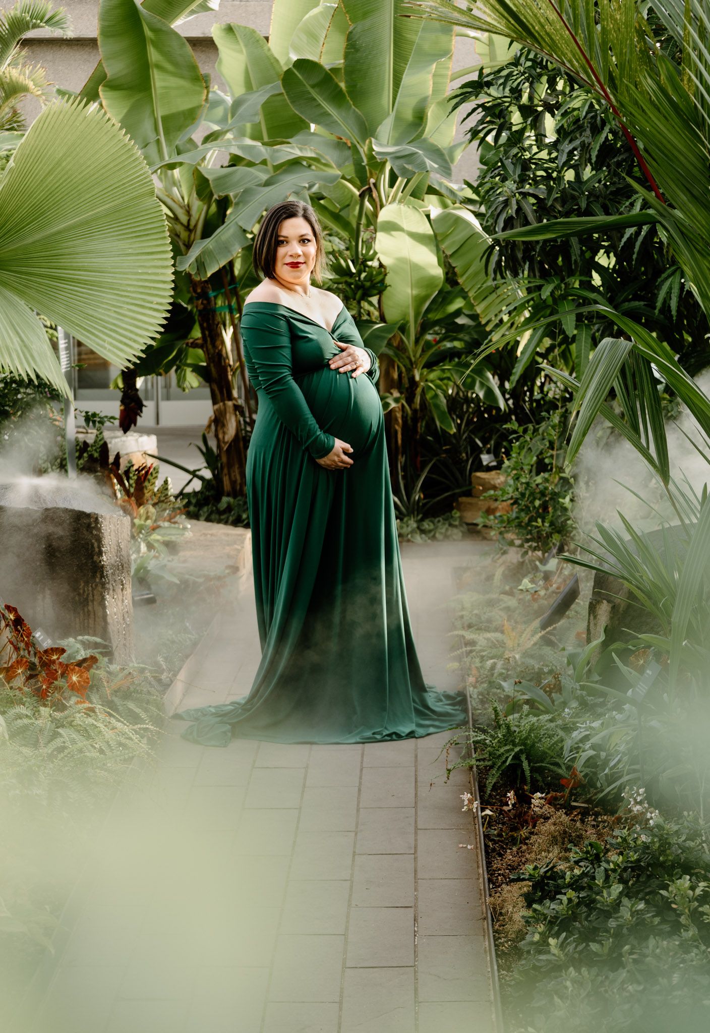 Expectant mother in flowing dark green maternity dress surrounded by tropical foliage and mist.