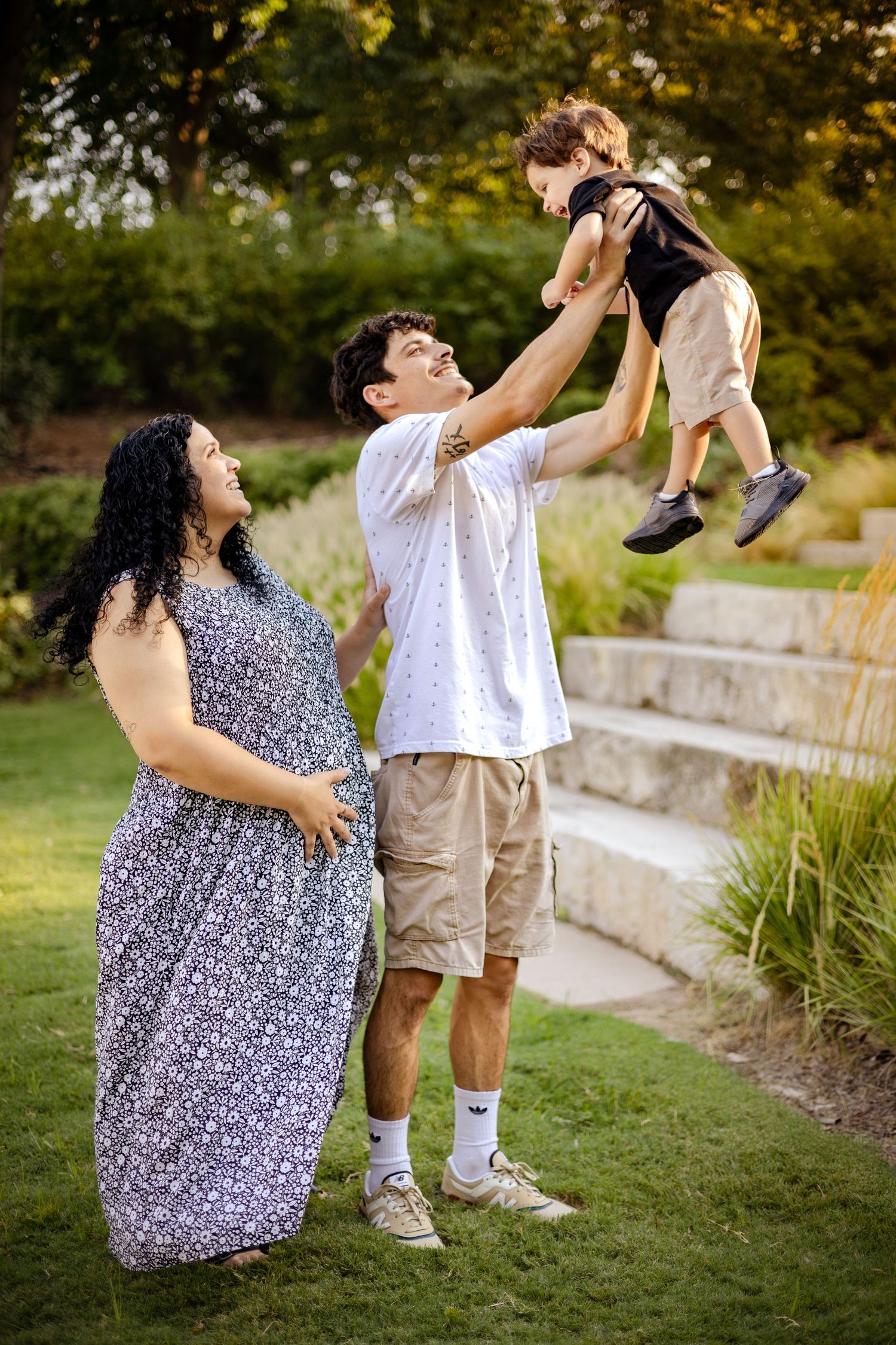 Happy family with pregnant mother and father lifting toddler in a park.
