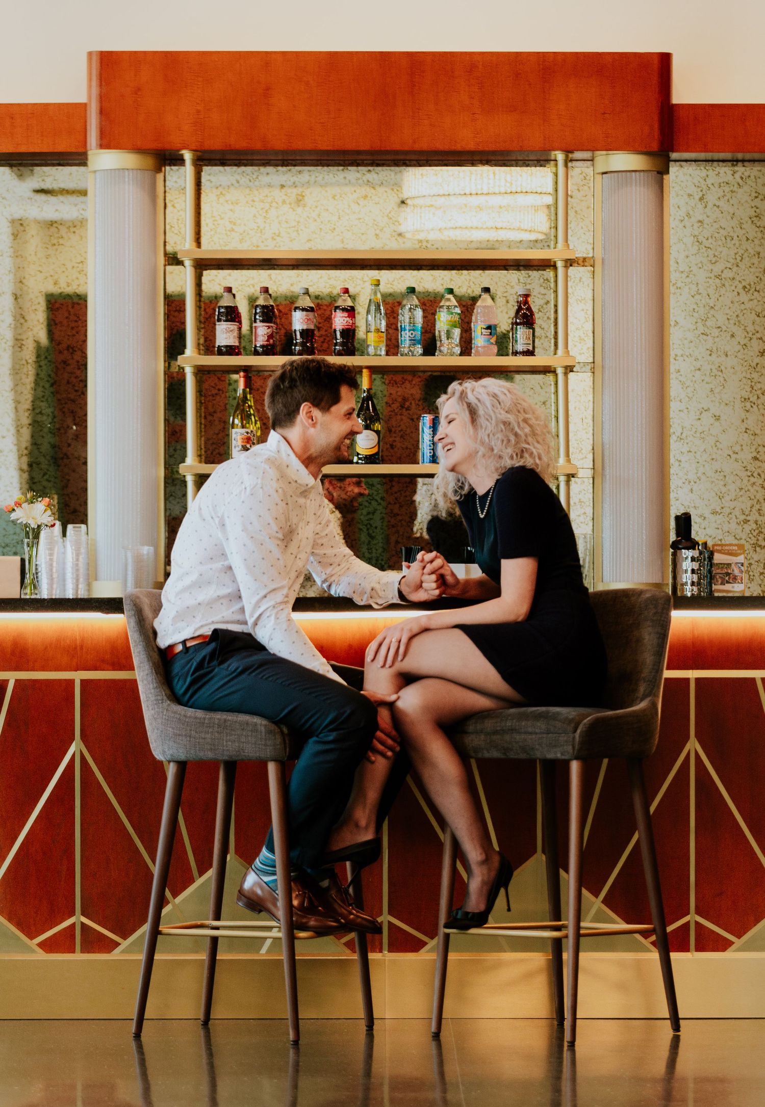 Couple laughing and holding hands at a stylish retro bar with gold-accented shelving behind them.