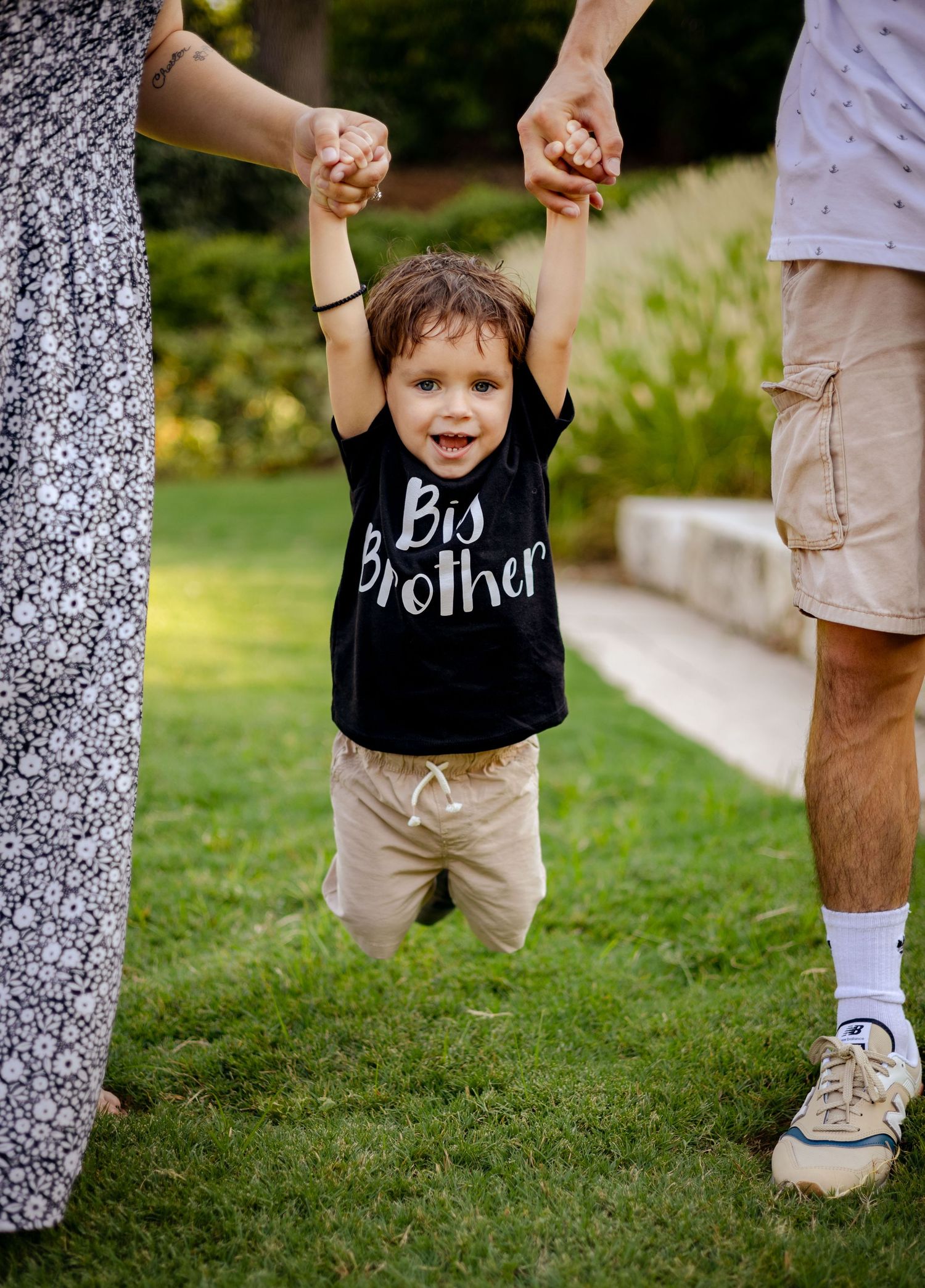 Smiling toddler in Big Brother shirt being lifted by parents hands in a park.