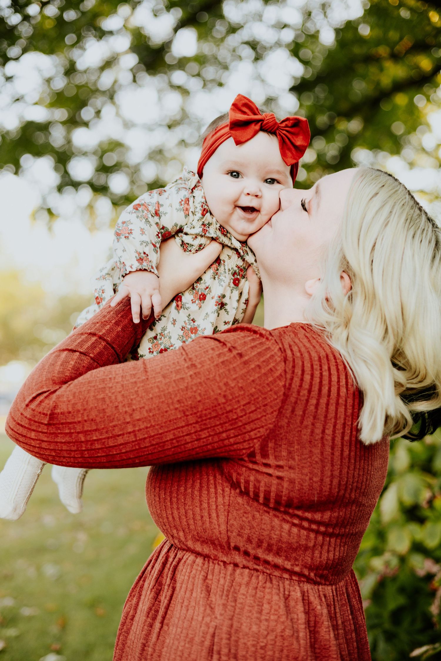 A blonde mother in a rust-red dress kisses her baby girl wearing a red bow headband outdoors.