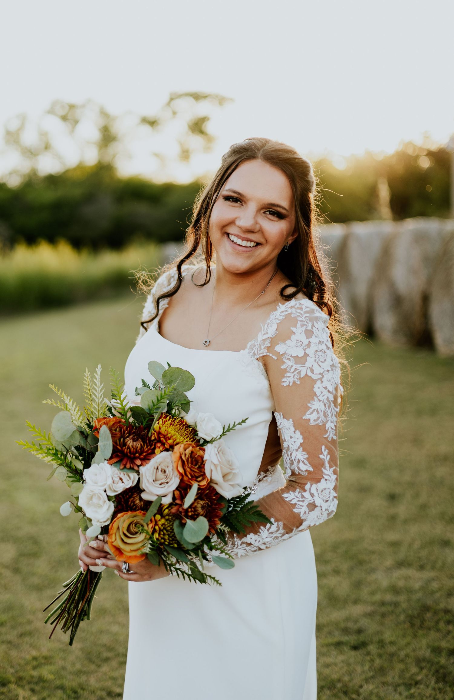 Smiling bride in lace sleeve wedding dress holding autumn bouquet at golden hour.