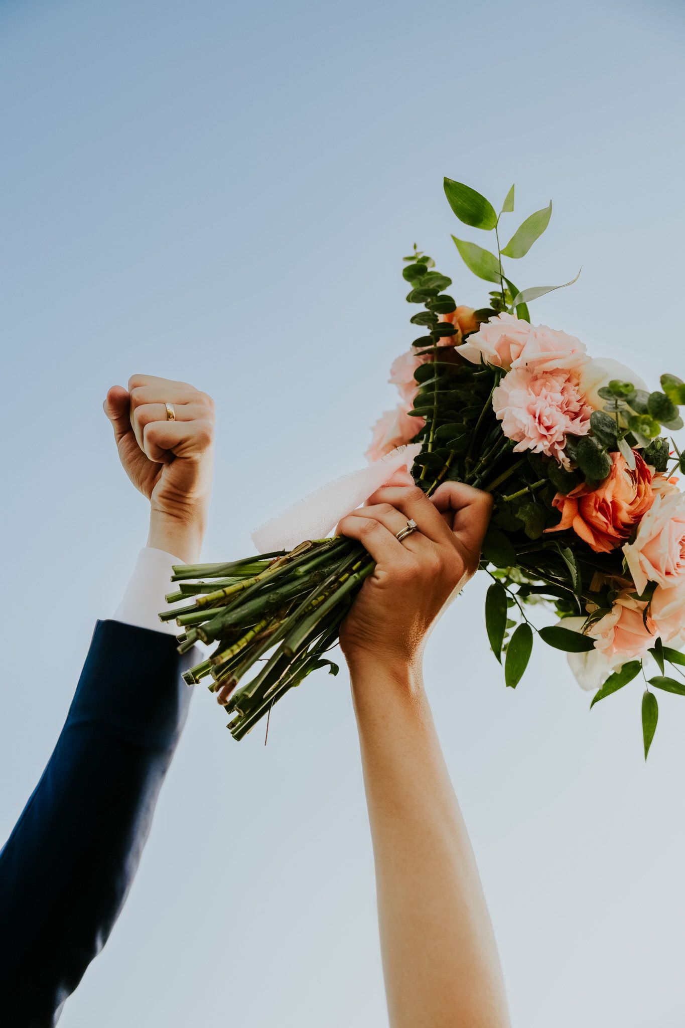 Newlyweds raise hands with wedding bouquet of pink roses against blue sky, showing wedding rings.