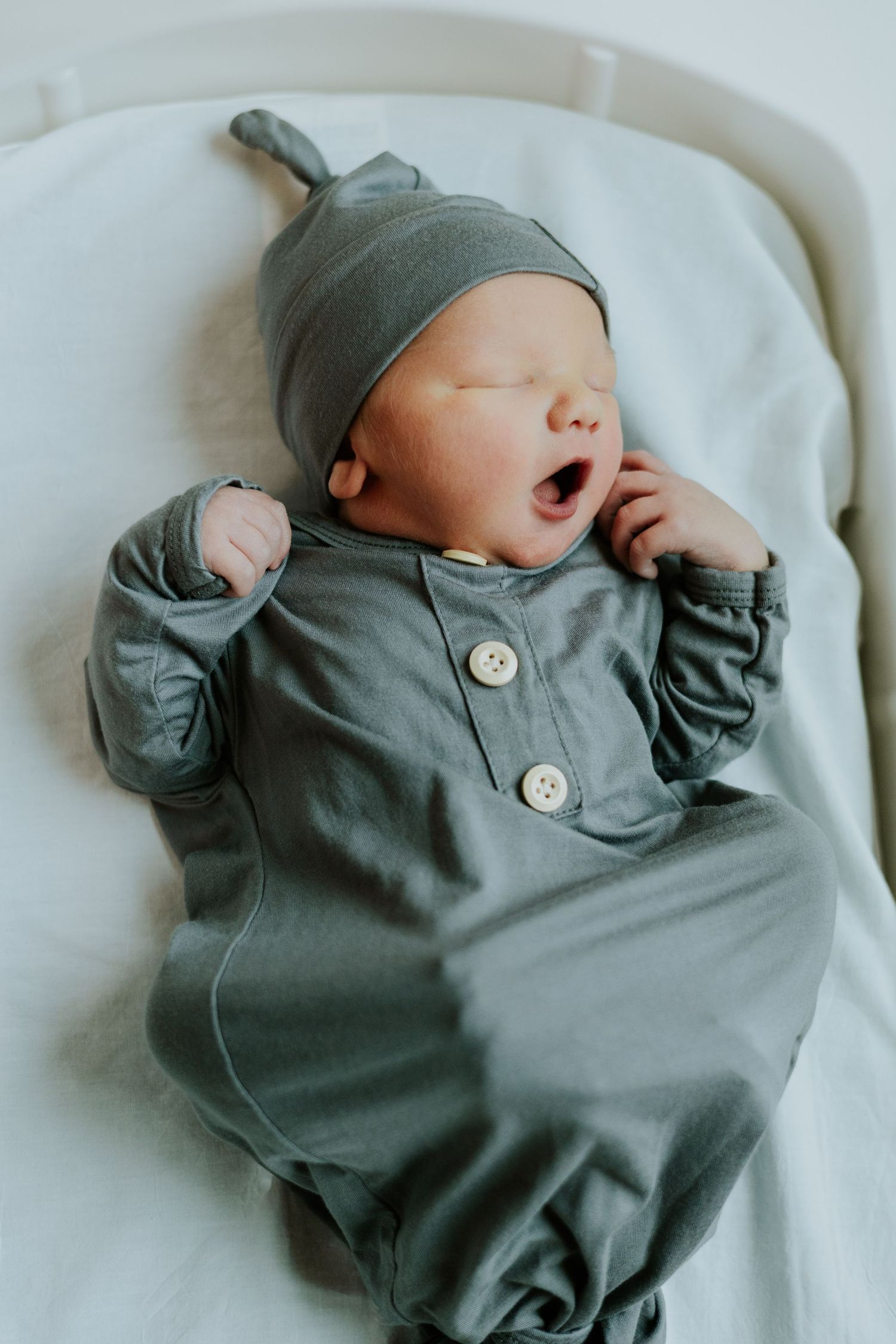 Newborn baby yawning in sage green knotted gown and matching hat lying on white bassinet.