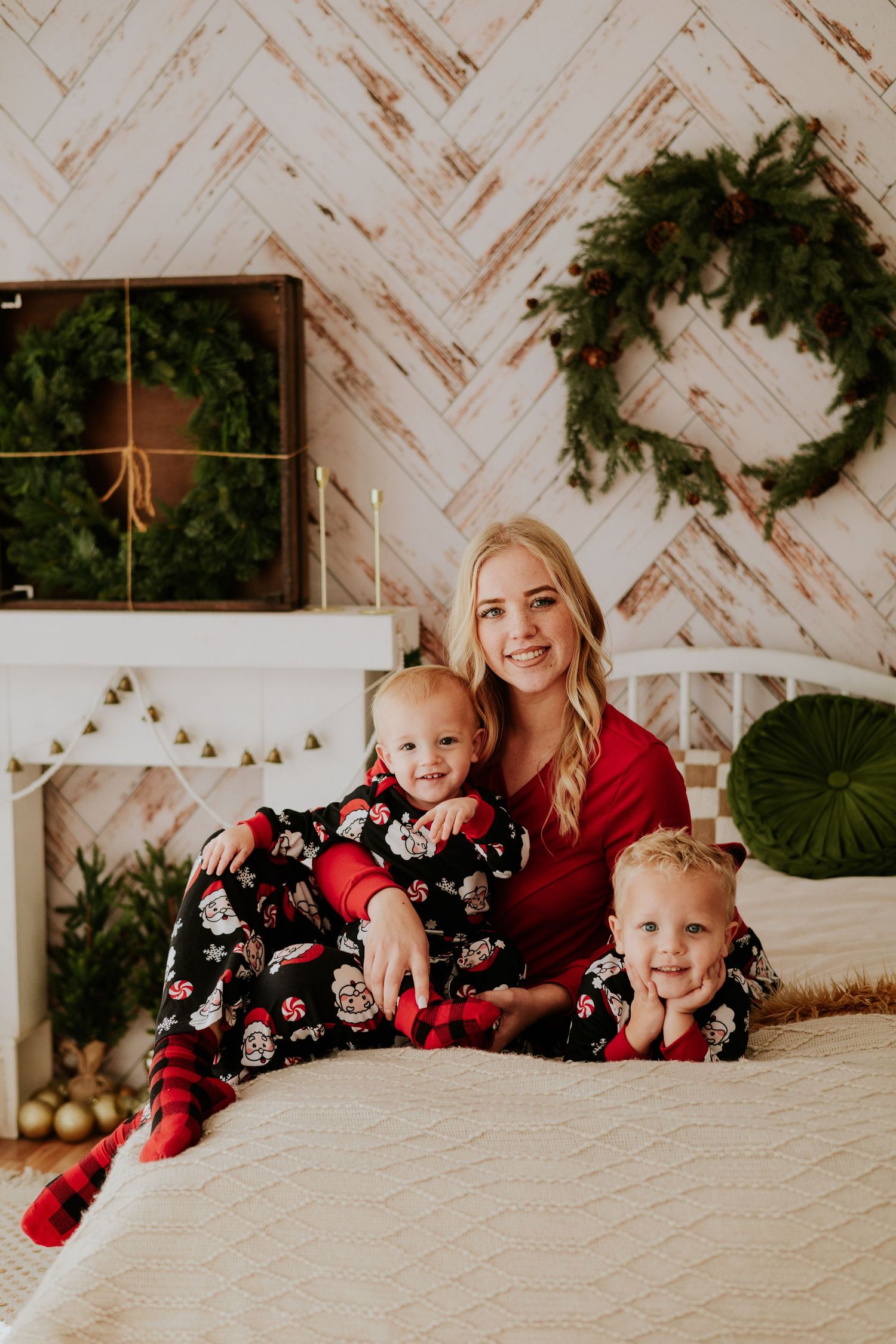 Mom and two young children in matching Christmas pajamas sitting on a bed with holiday decorations.