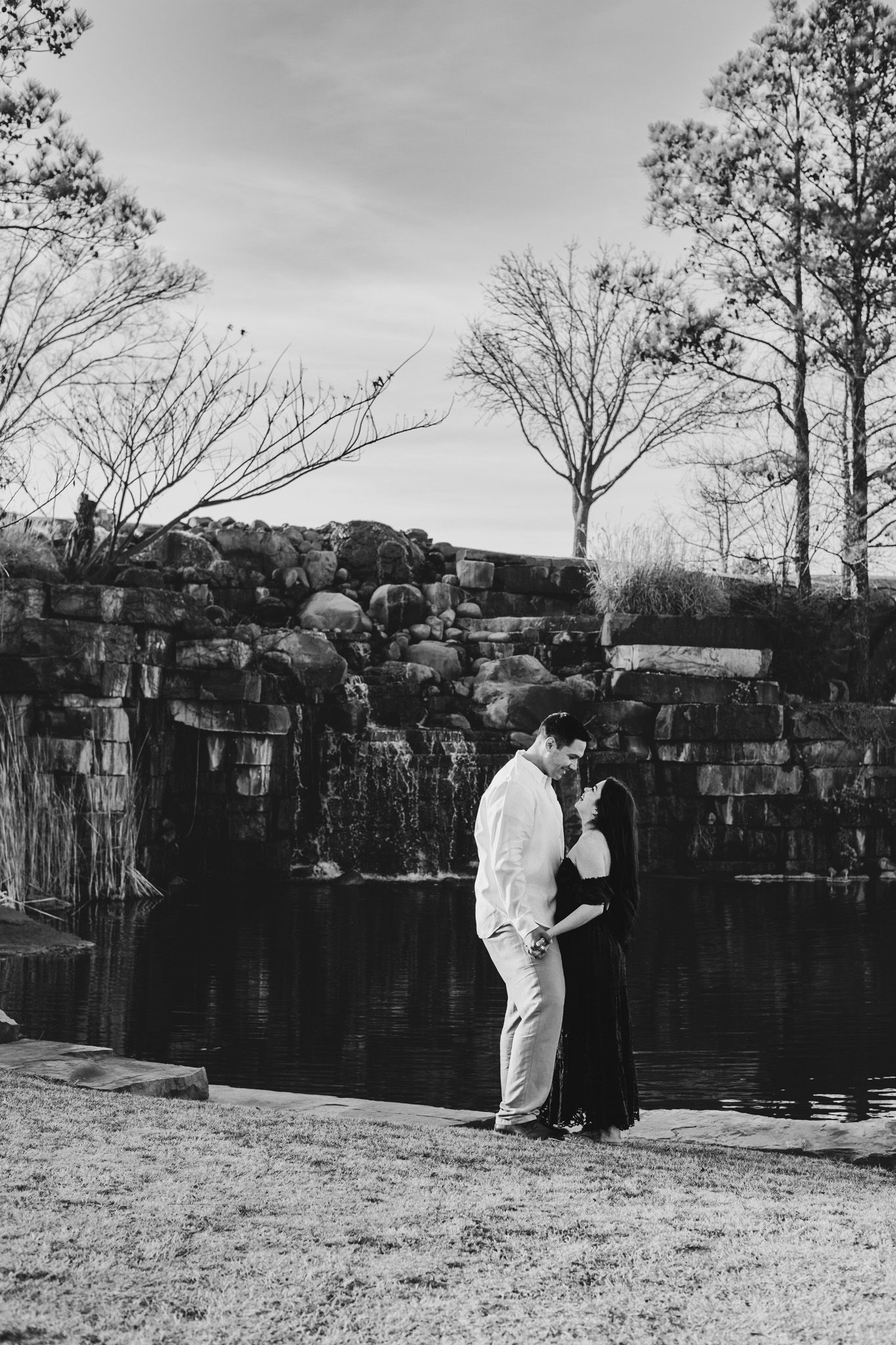 Black and white image of a romantic couple embracing near a stone waterfall outdoors.