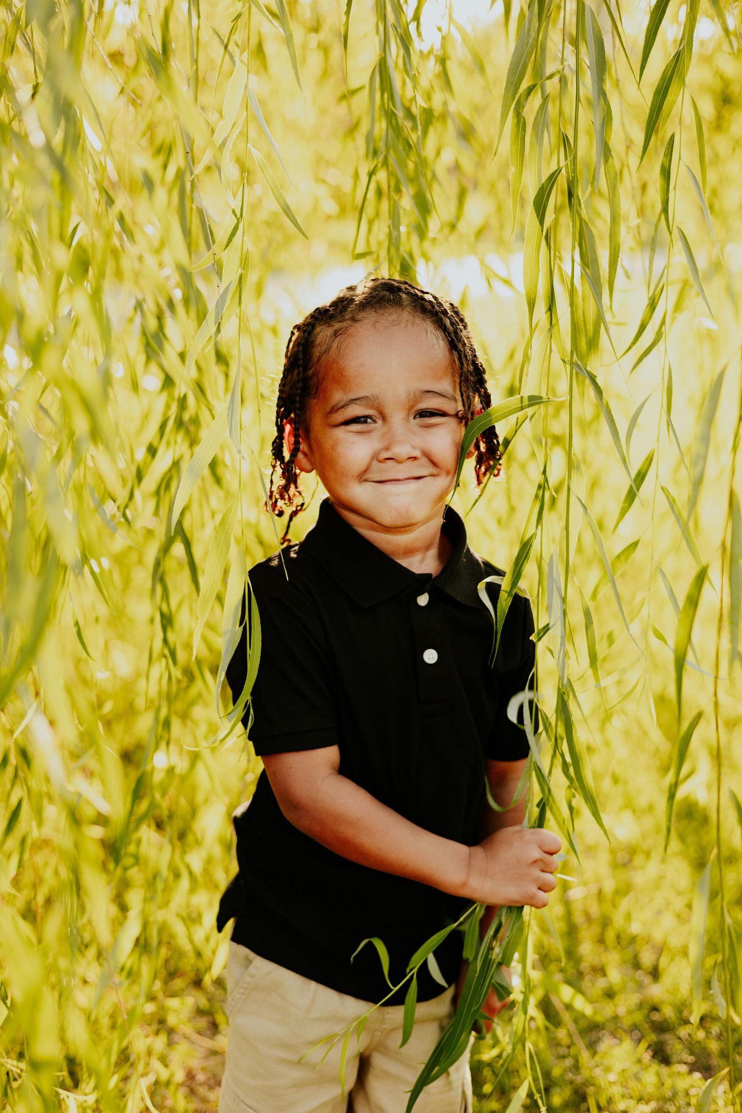 Smiling young boy with braids in black polo shirt standing among weeping willow branches outdoors.