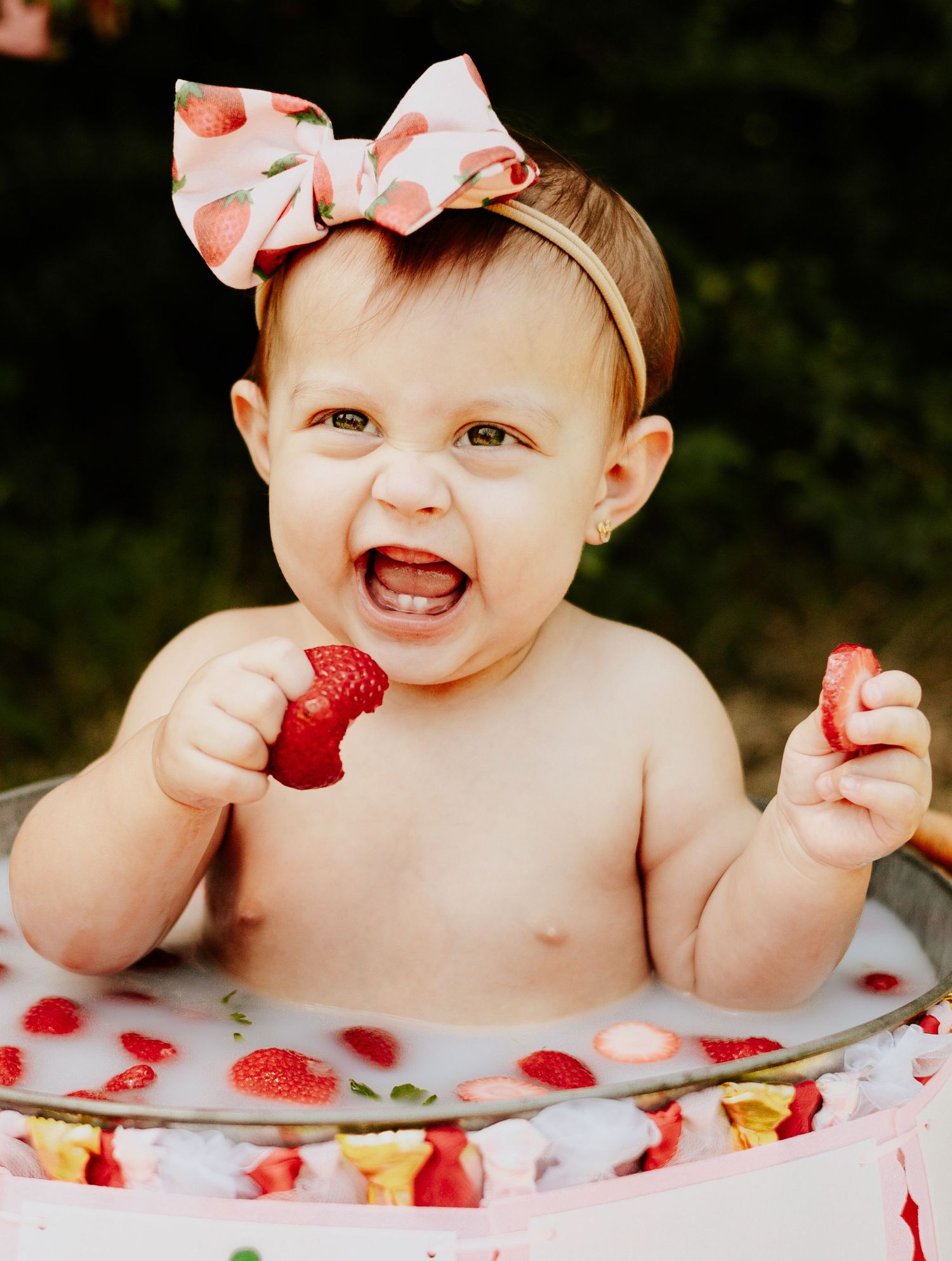 A happy baby girl with a strawberry bow headband sits in a milk bath holding fresh strawberries.