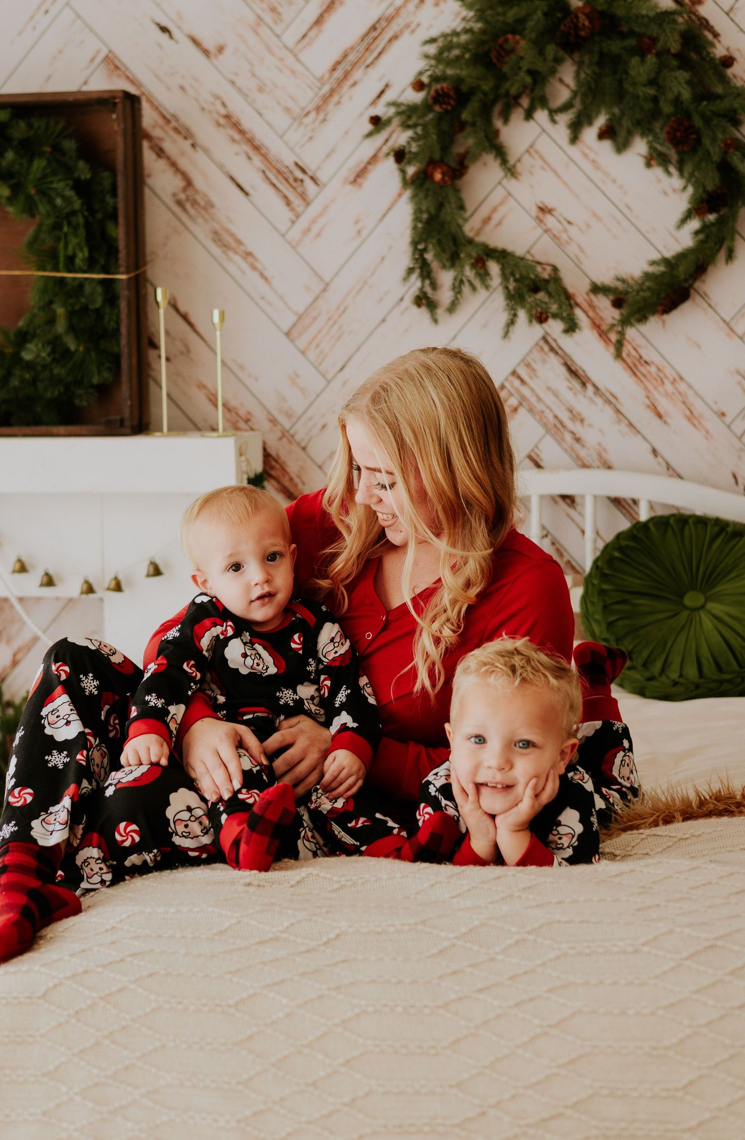 Blonde mother in red pajamas sitting on bed with two young children in Christmas holiday pajamas.