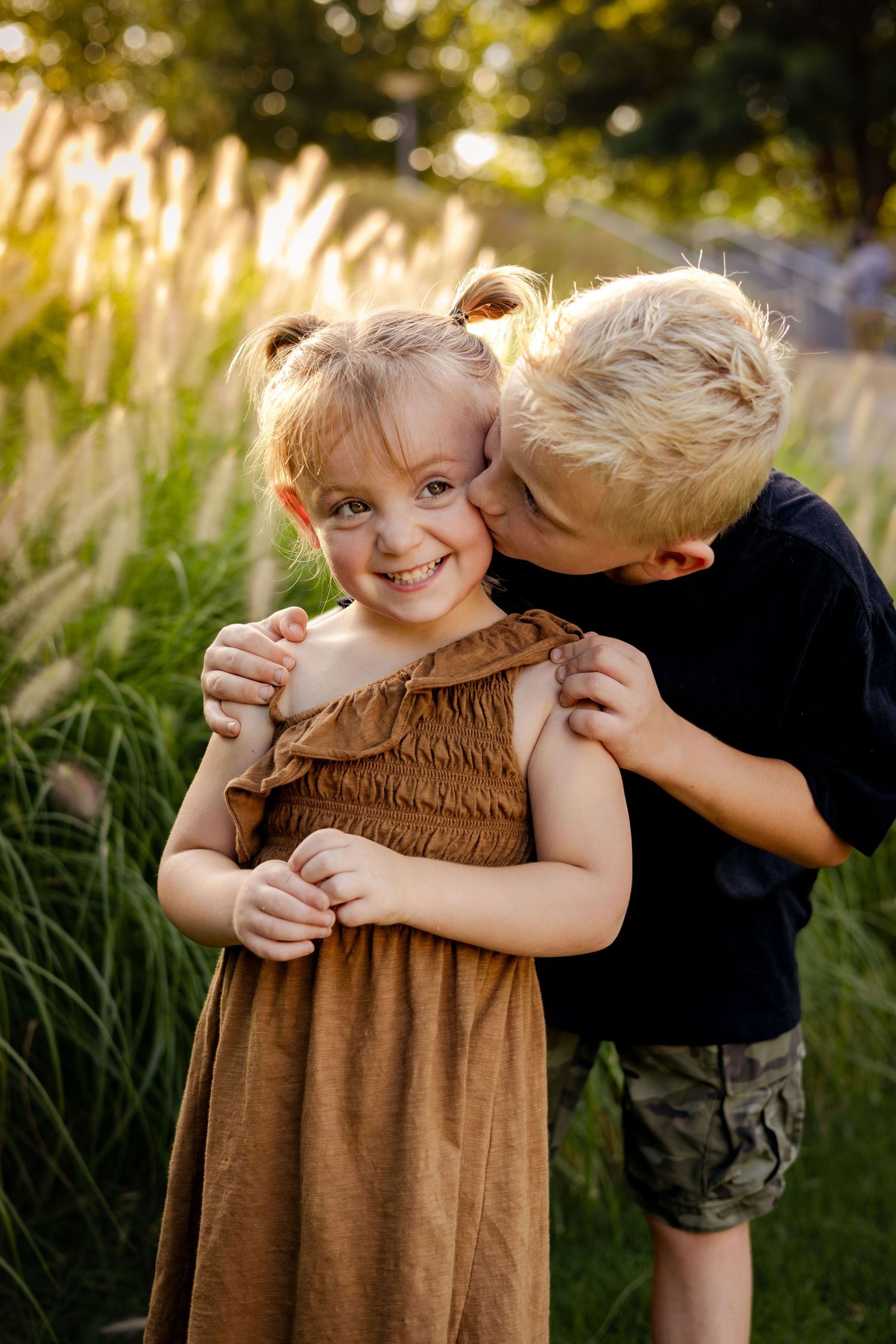 Blonde boy in black shirt kissing smiling little sister in brown dress outdoors in golden sunset light.