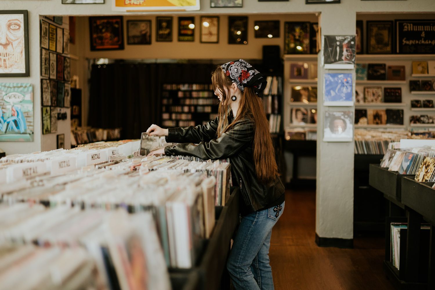 Woman browsing vinyl records in a vintage music store, wearing a bandana and leather jacket.
