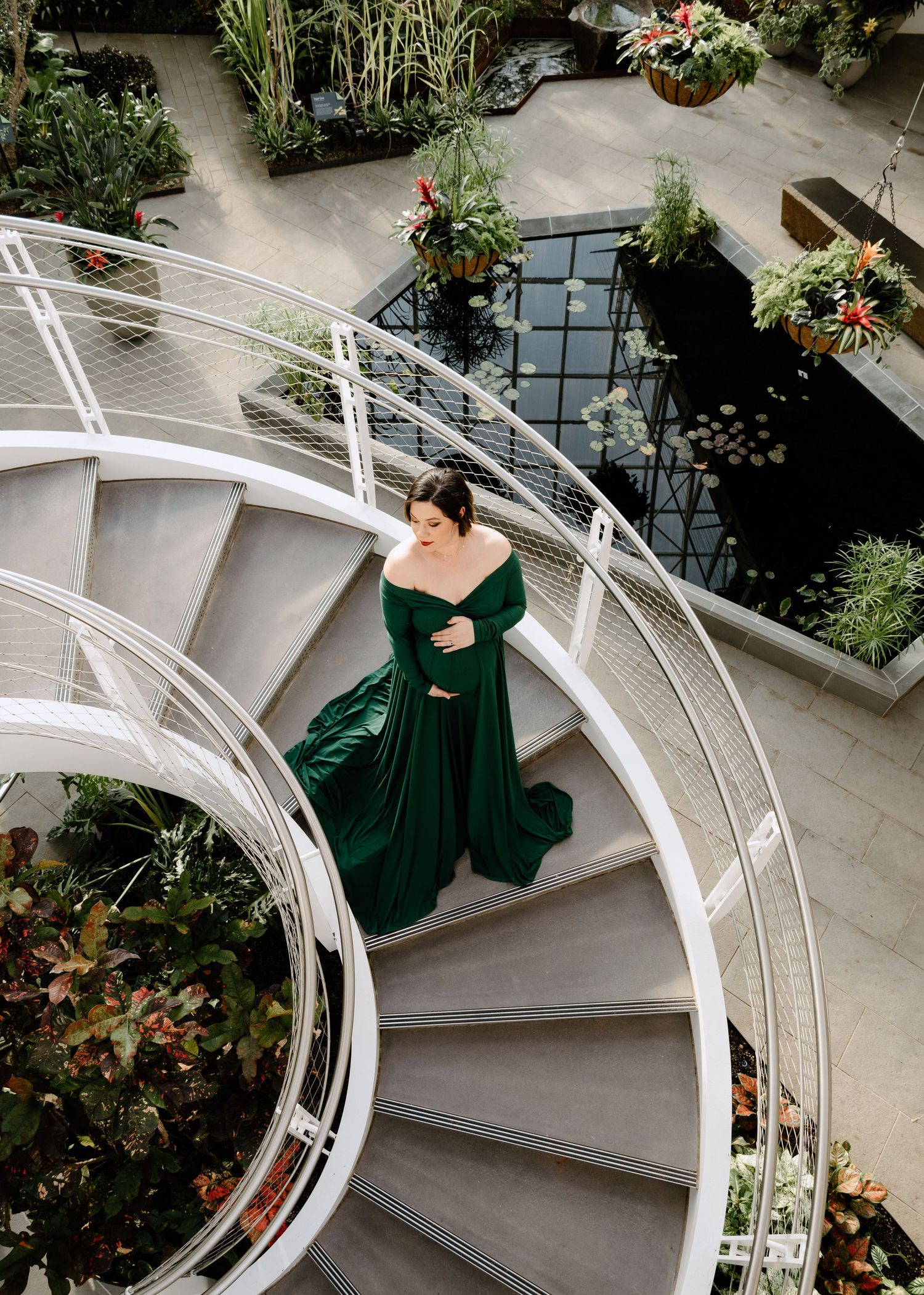 Woman in emerald green gown poses gracefully on a white spiral staircase surrounded by lush indoor plants.