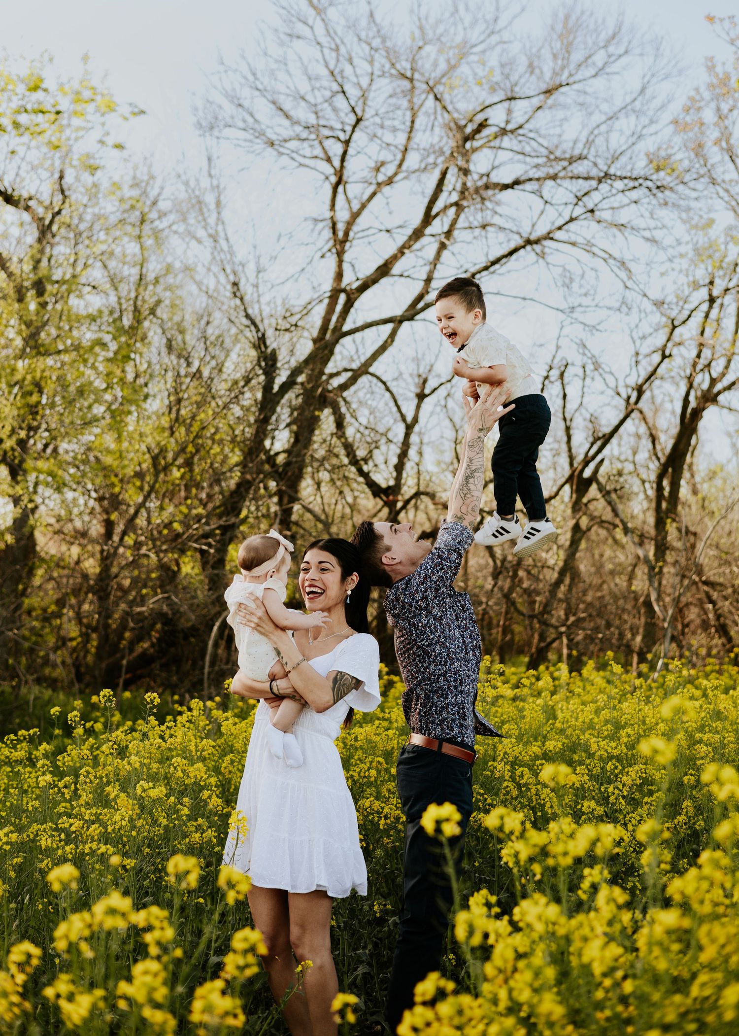 Family laughing in yellow wildflower field, dad lifting toddler overhead while mom holds baby on sunny spring day.