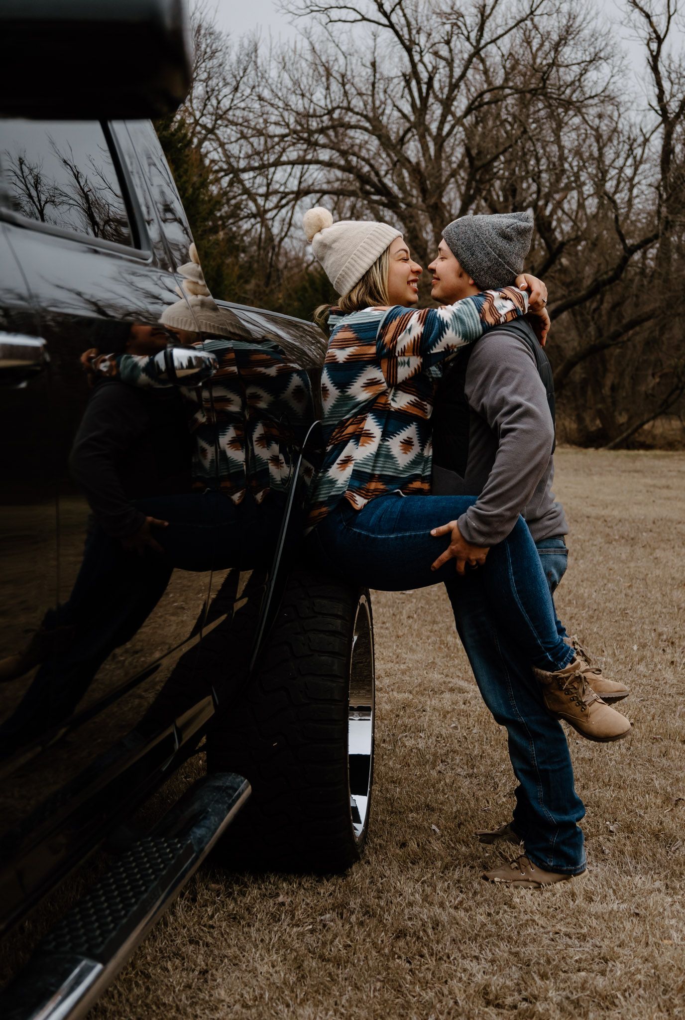 Couple sharing a tender moment leaning against a black lifted truck outdoors in winter.