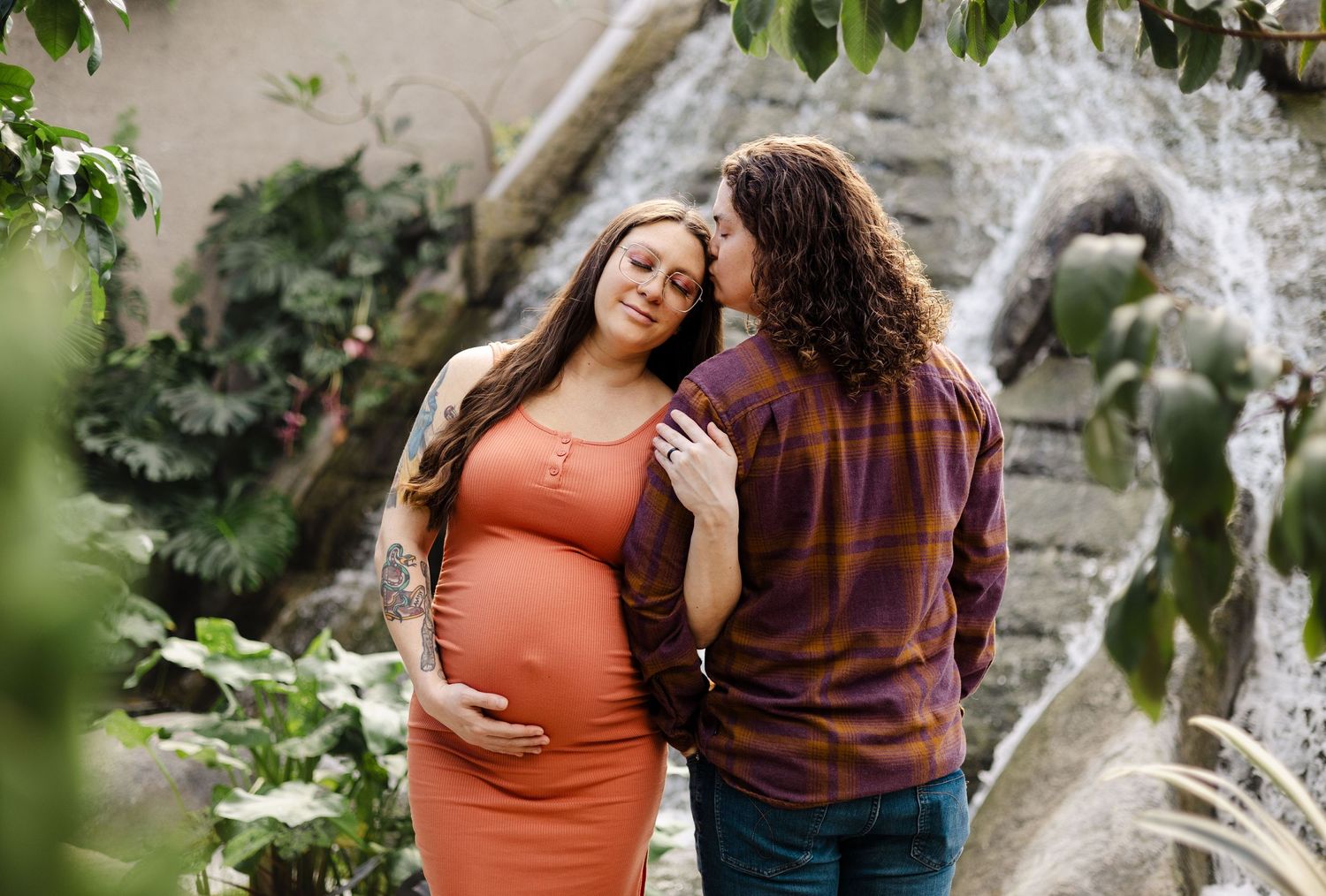 Pregnant woman in orange dress with partner sharing a tender moment near a lush garden waterfall.