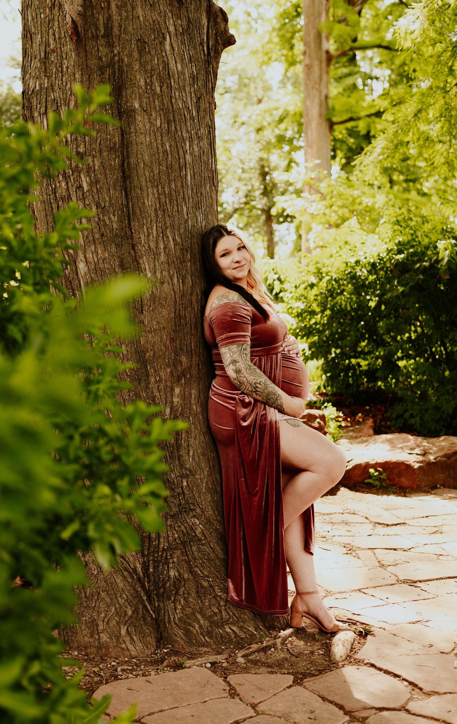 Pregnant woman in burgundy velvet maternity dress leaning against tree trunk in lush green garden setting.