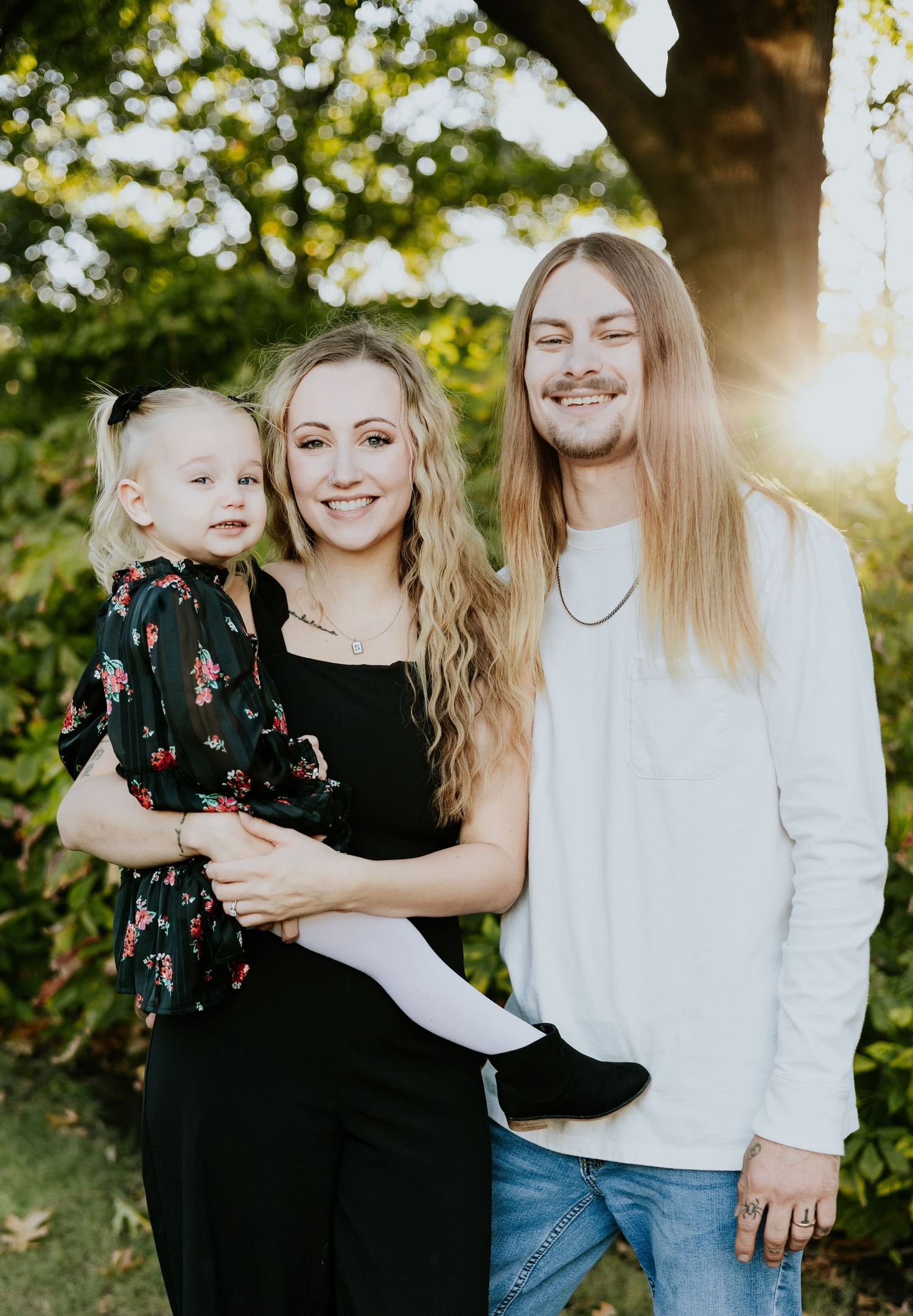 A smiling family of three poses outdoors at golden hour, with a woman holding a toddler girl beside a long-haired man.