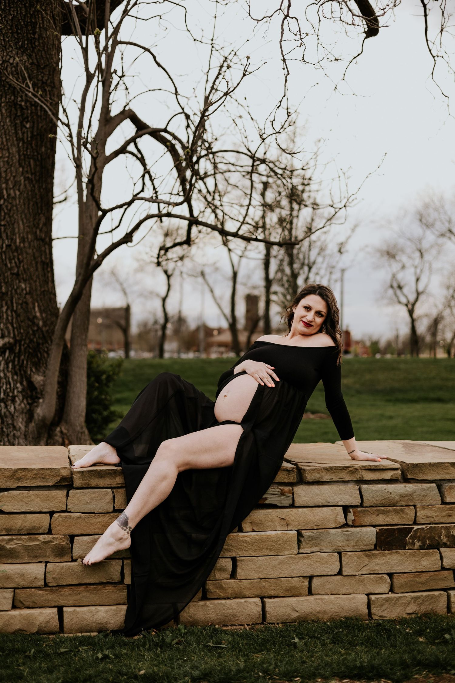 Pregnant woman in black maternity gown reclines on stone wall in park during golden hour.