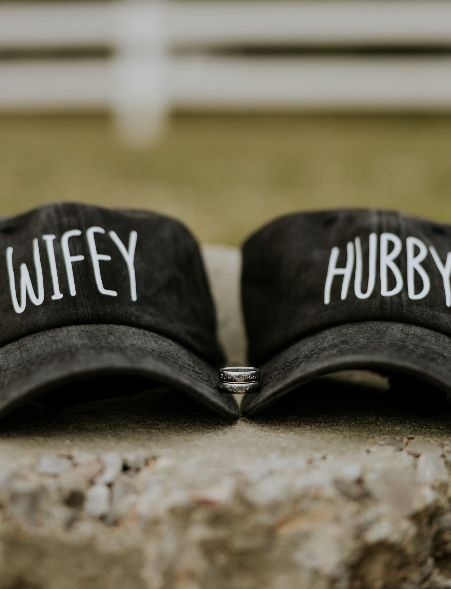 Two black baseball caps labeled Wifey and Hubby with wedding rings placed between them on stone surface.