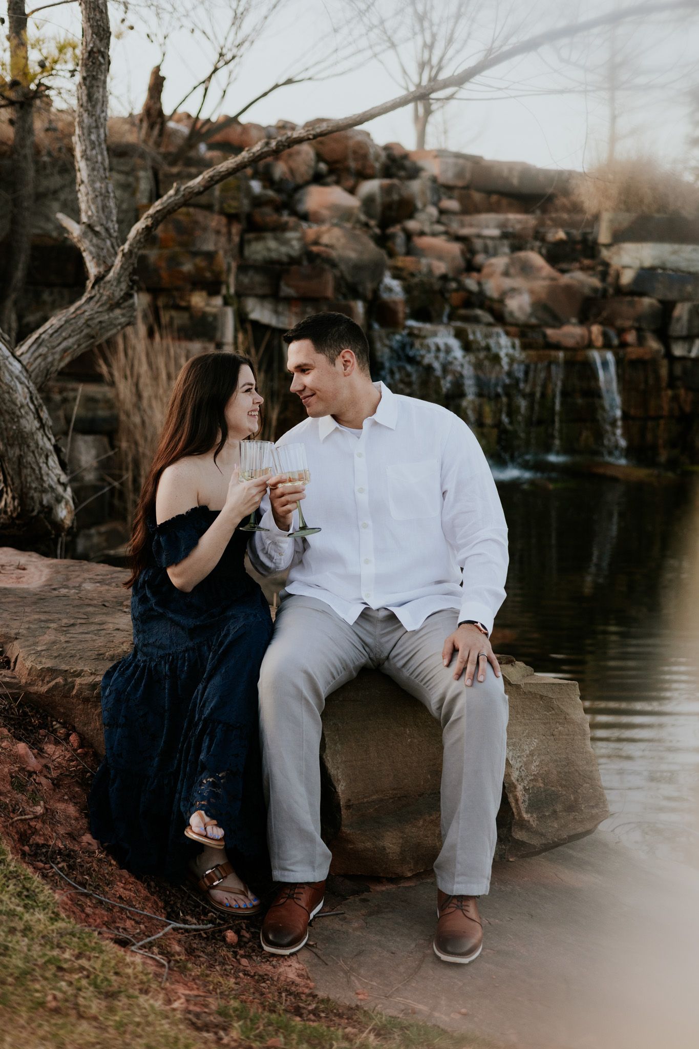 Happy engaged couple sitting on rocks by waterfall toasting with champagne glasses in nature setting.