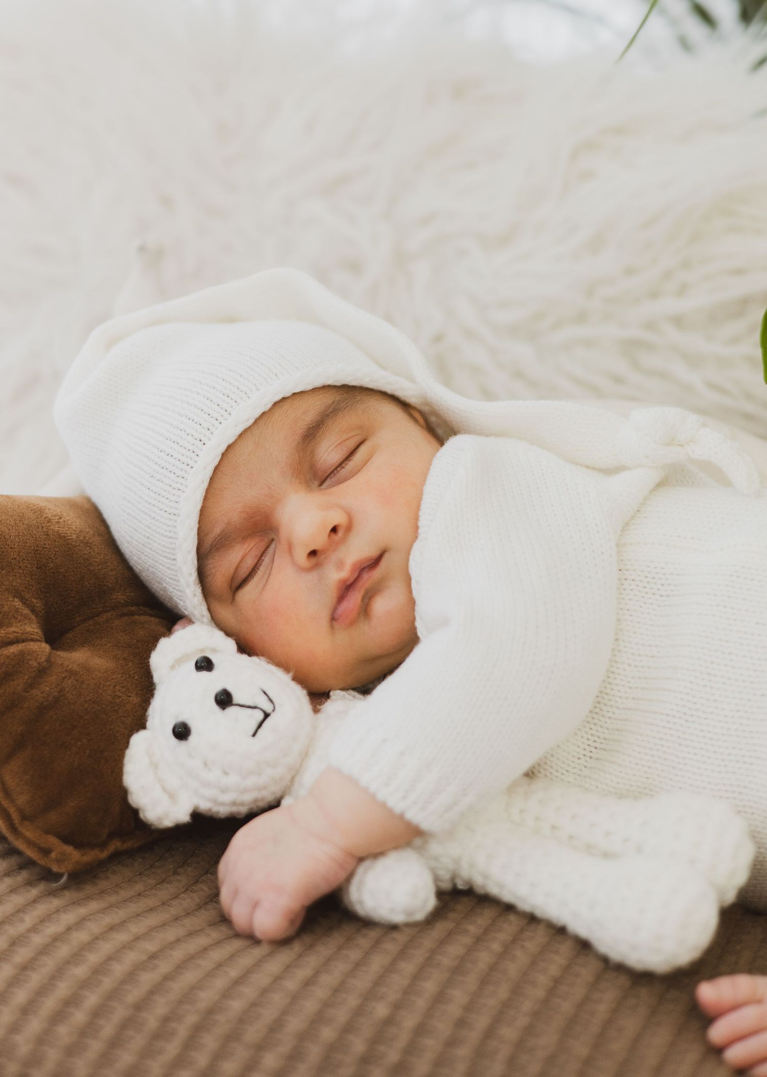 Sleeping newborn in white knit outfit and hat cuddling small crocheted teddy bear on brown pillow.