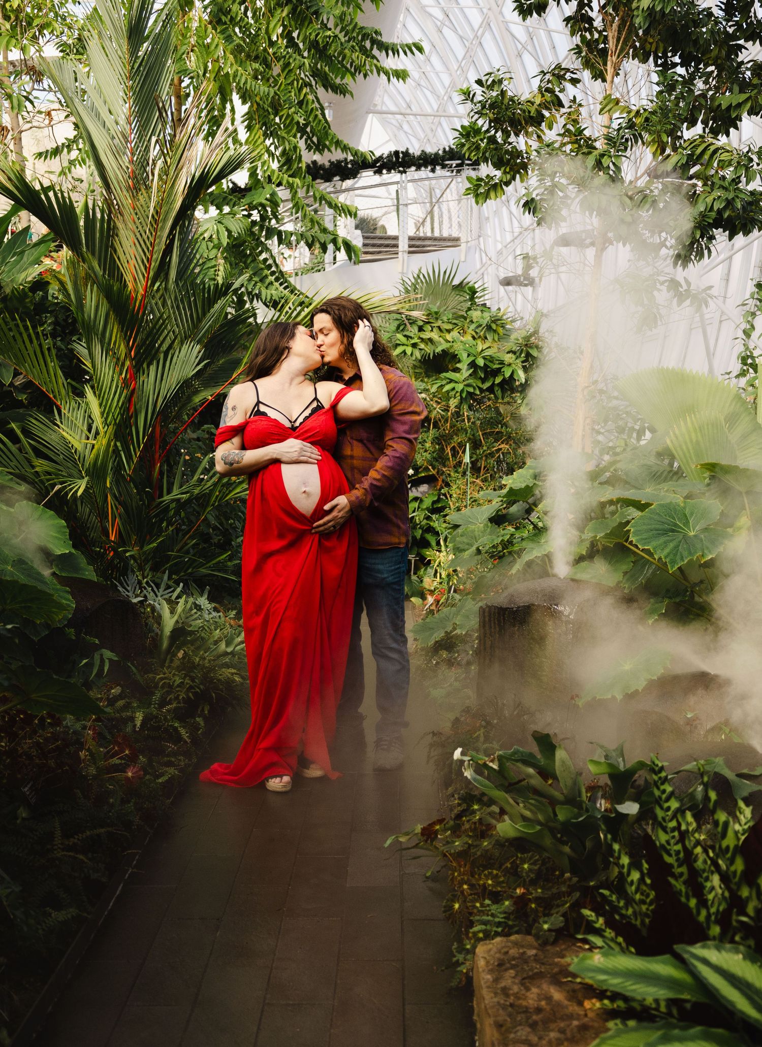 Couple embracing in a misty tropical greenhouse during a stunning red dress maternity shoot.