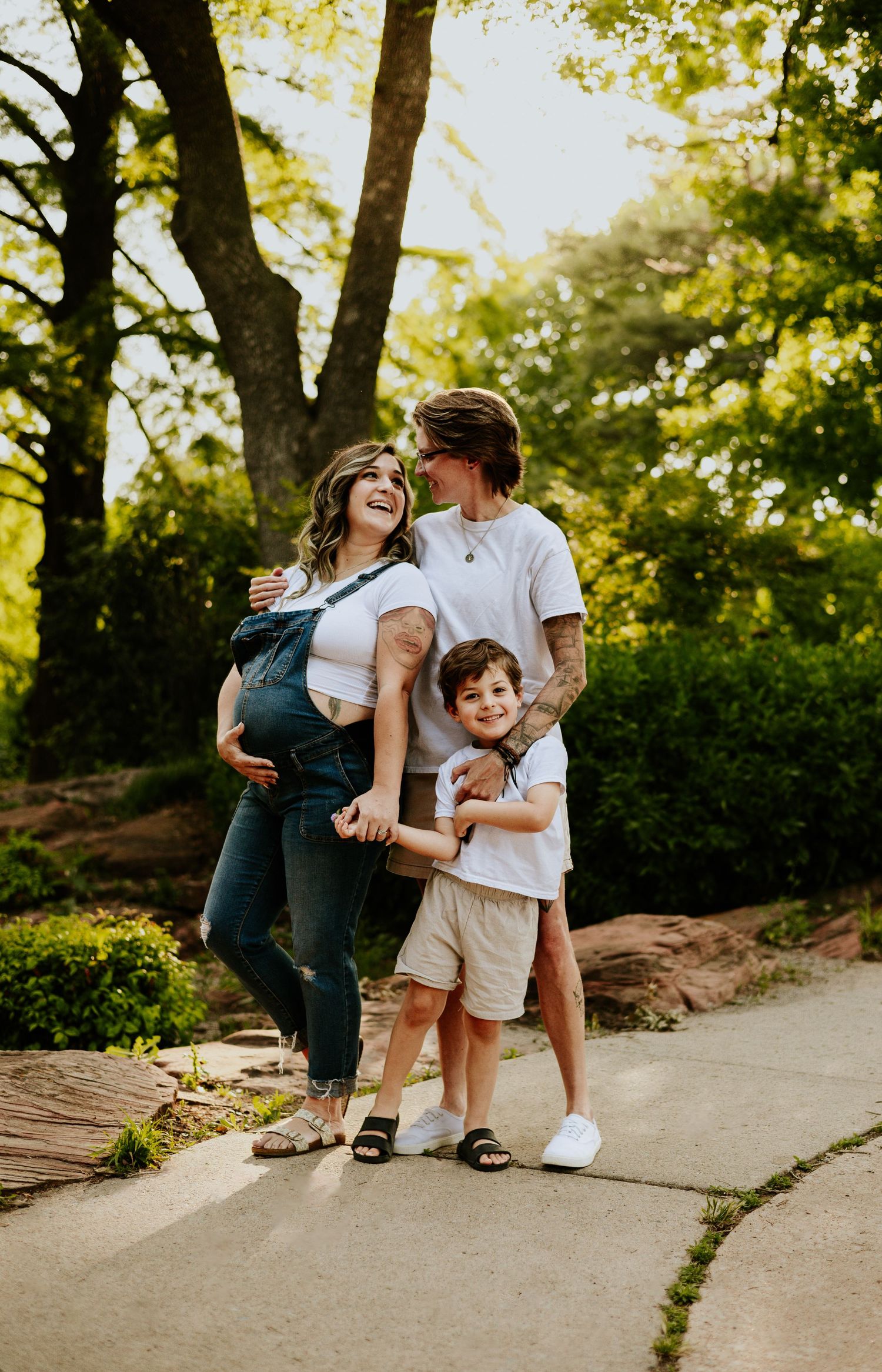 A happy pregnant woman, her partner, and young son share a joyful moment on a sunny tree-lined path.